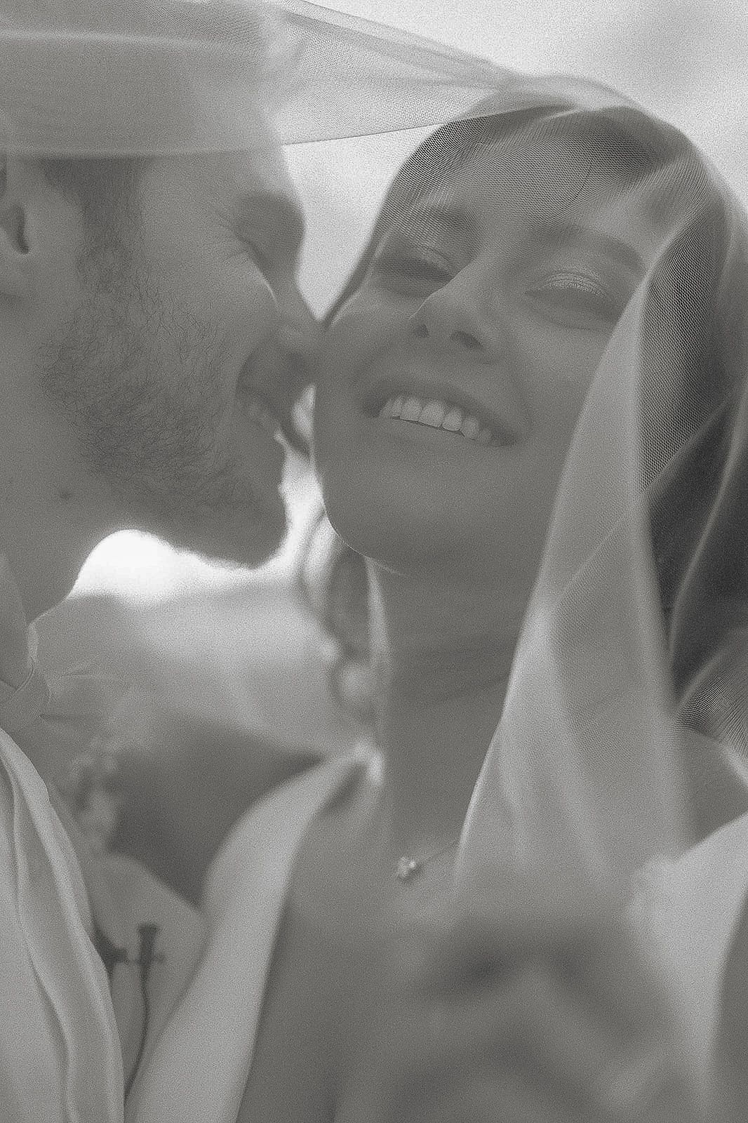 Black and white wedding photo of smiling bride under veil as groom leans in for a kiss.