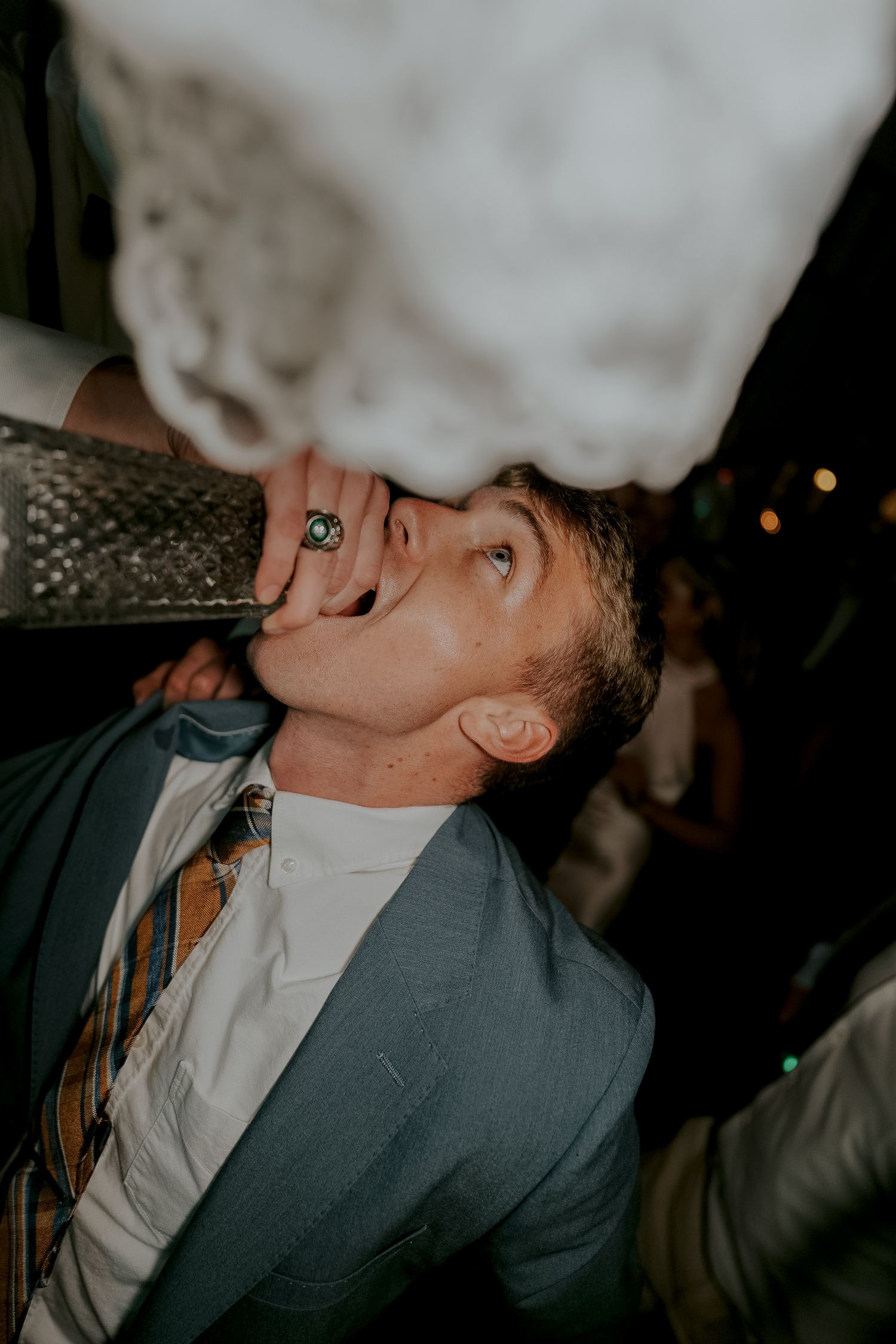 Wedding guest in blue suit drinking from bottle at lively reception party.