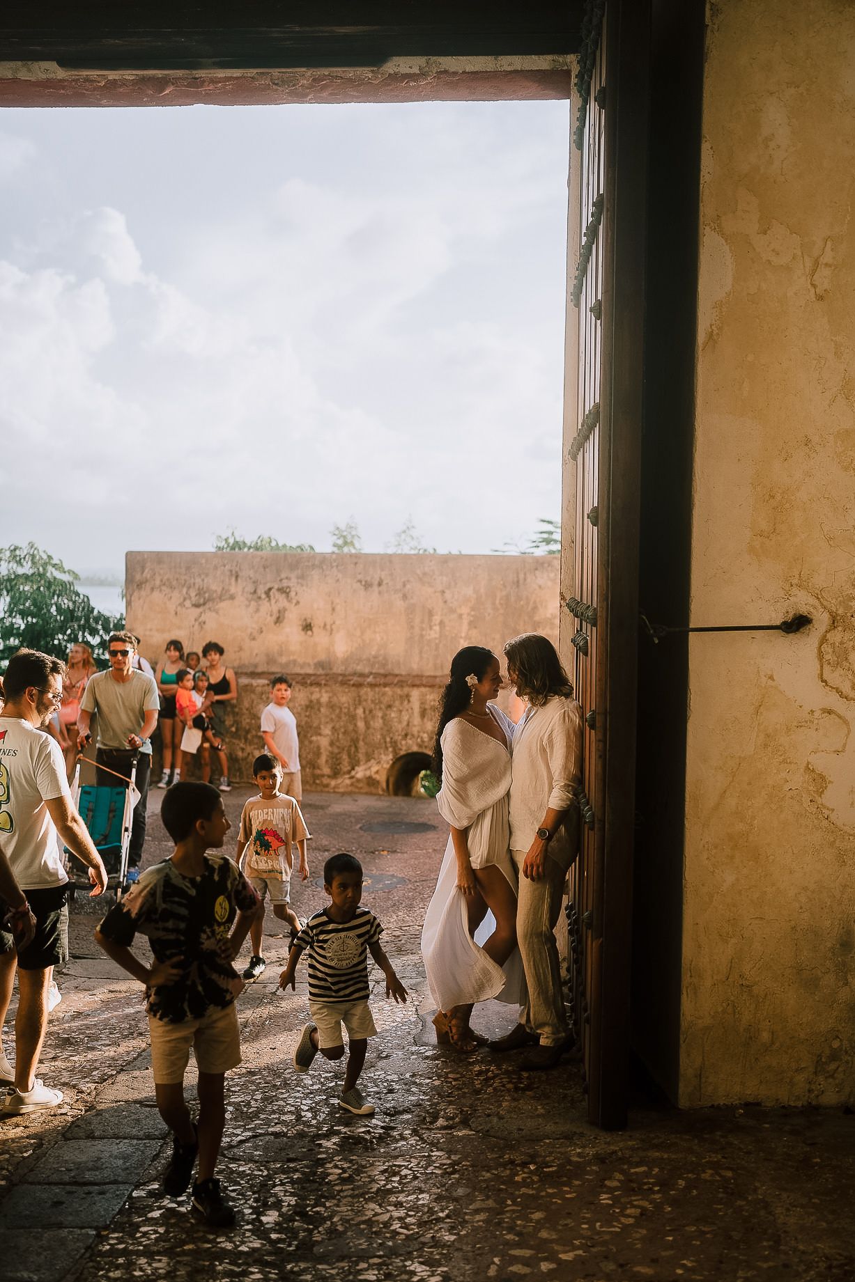 Couple shares a kiss at historic stone fortress entrance surrounded by tourists and children.