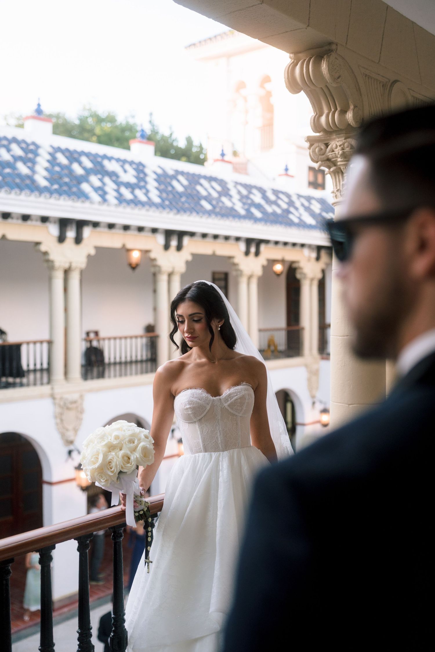 Bride in strapless white gown holds bouquet on balcony as groom in navy suit watches nearby.
