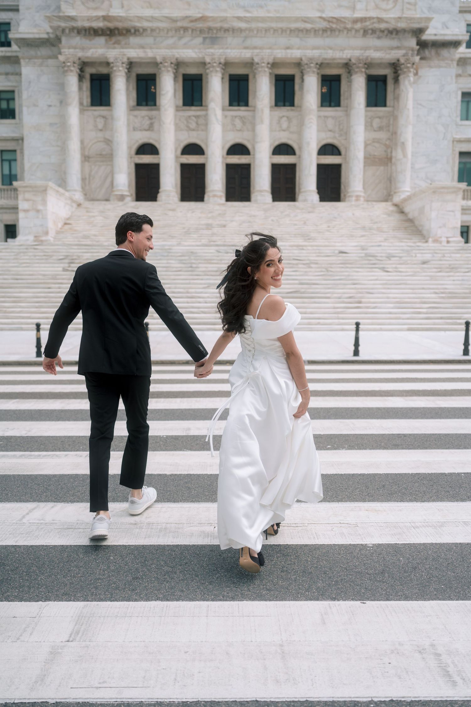 Bride in white off-shoulder gown and groom in black suit hold hands crossing street before grand marble building.