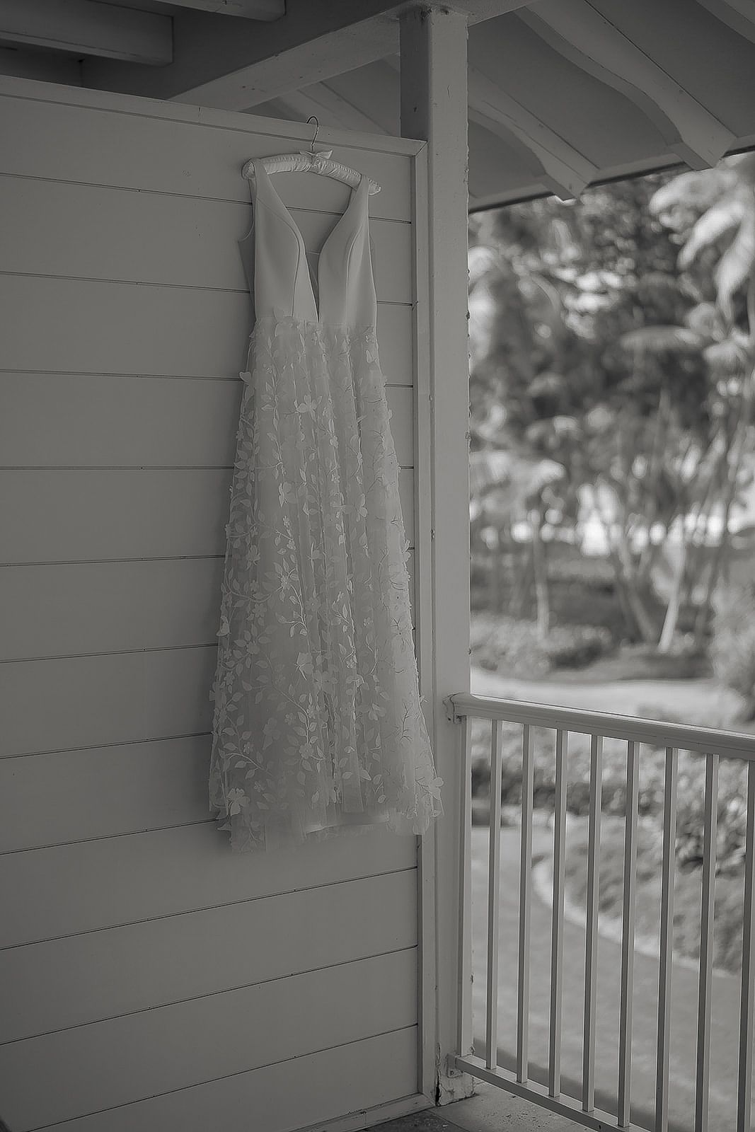 Black and white photo of a lace wedding dress hanging on a porch wall beside a balcony railing with trees in background.