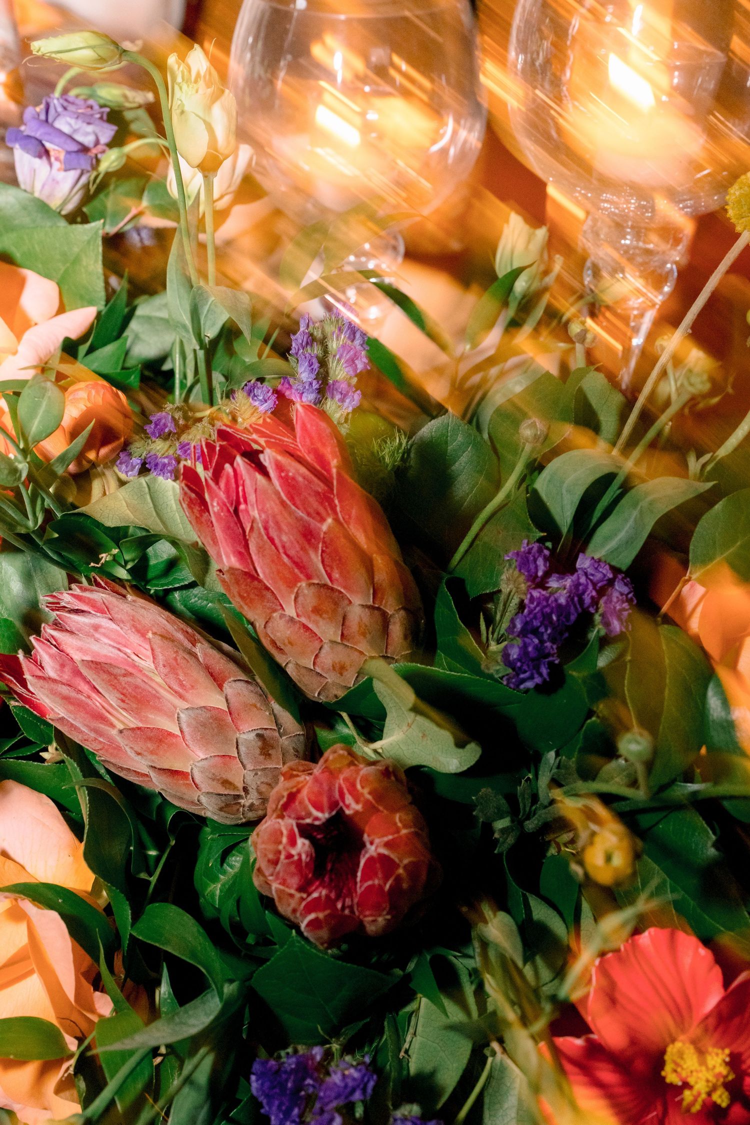 Colorful wedding floral arrangement featuring protea, roses, and purple flowers with glowing candles.