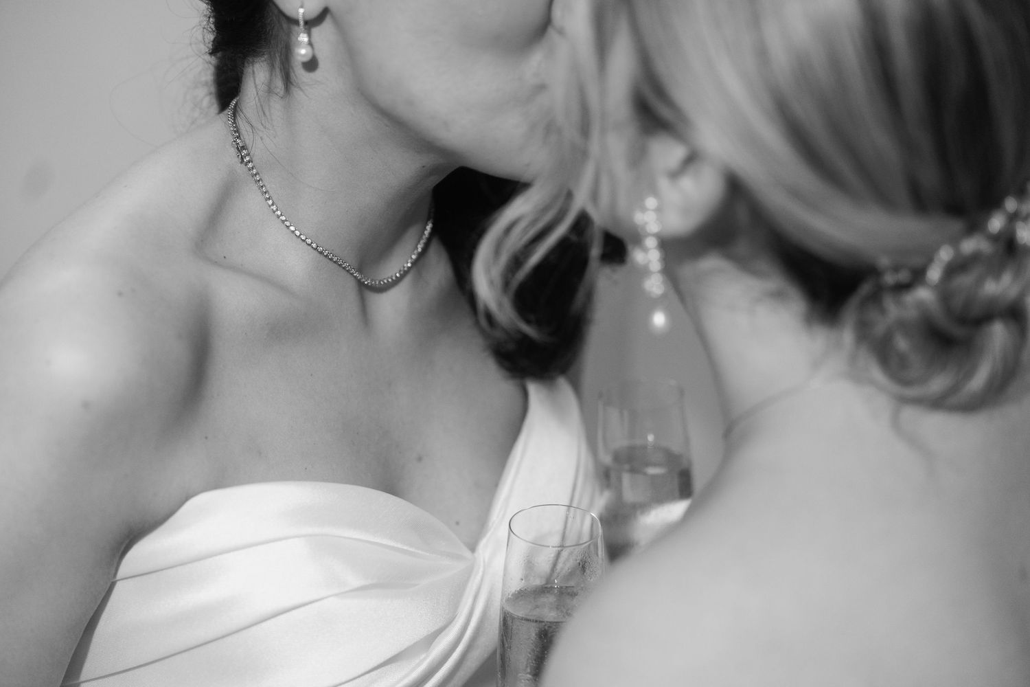 Two brides sharing a kiss in black and white wedding photo, one holding champagne glass.