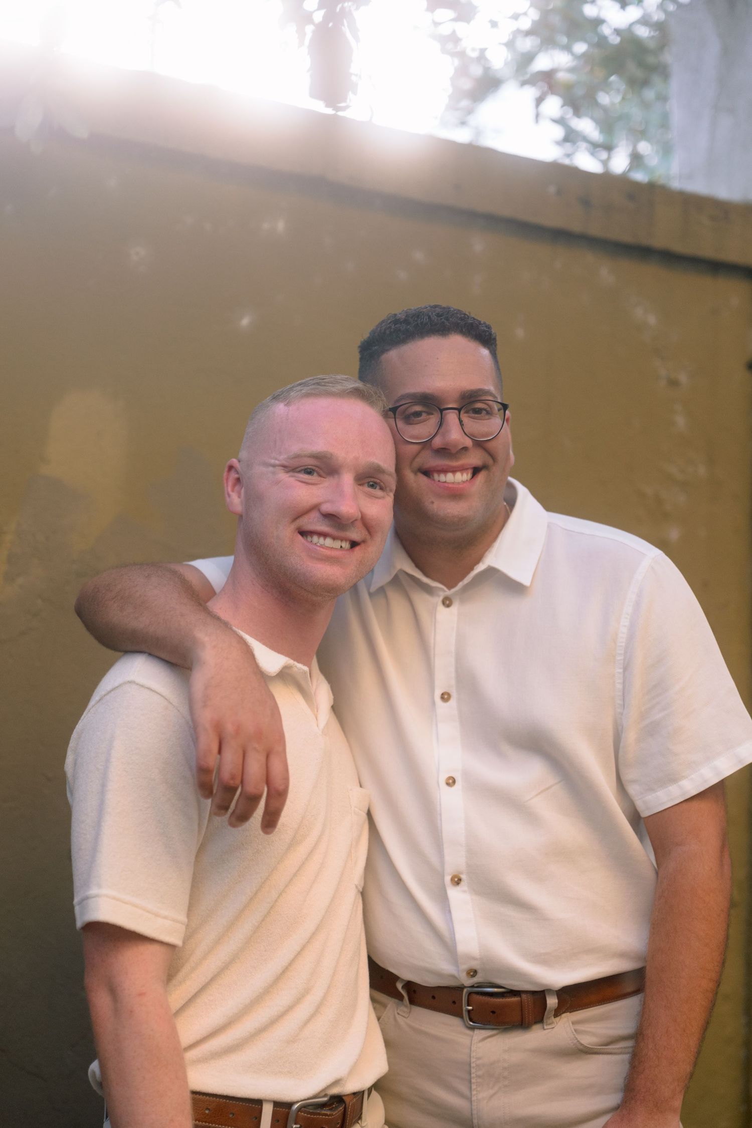 Two smiling men in white polo shirts pose together outdoors in warm sunlight.