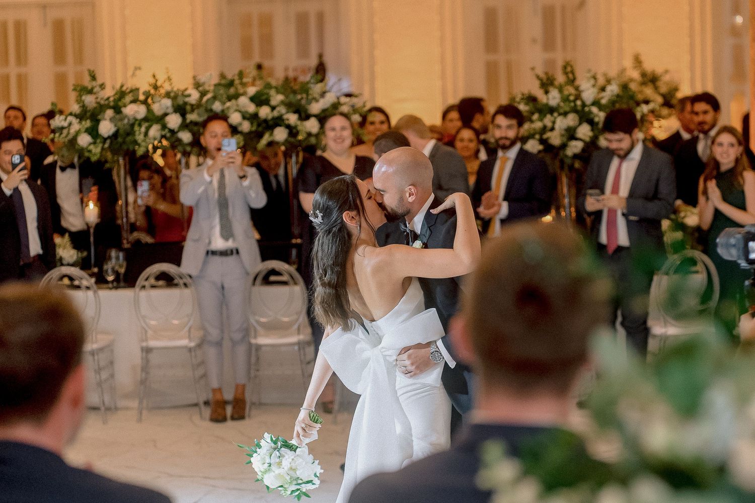 Bride and groom share first dance kiss surrounded by wedding guests in elegant white floral ballroom.