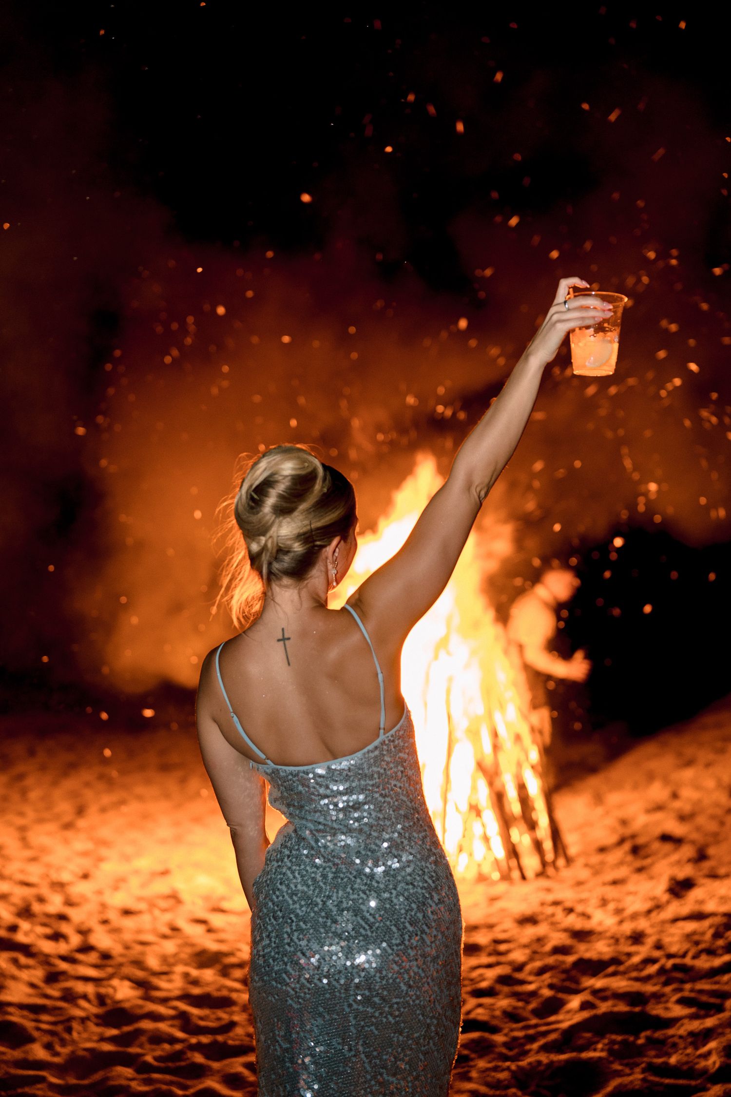 Woman in sequin dress raising a drink by a bonfire on the beach at night, with sparks flying.