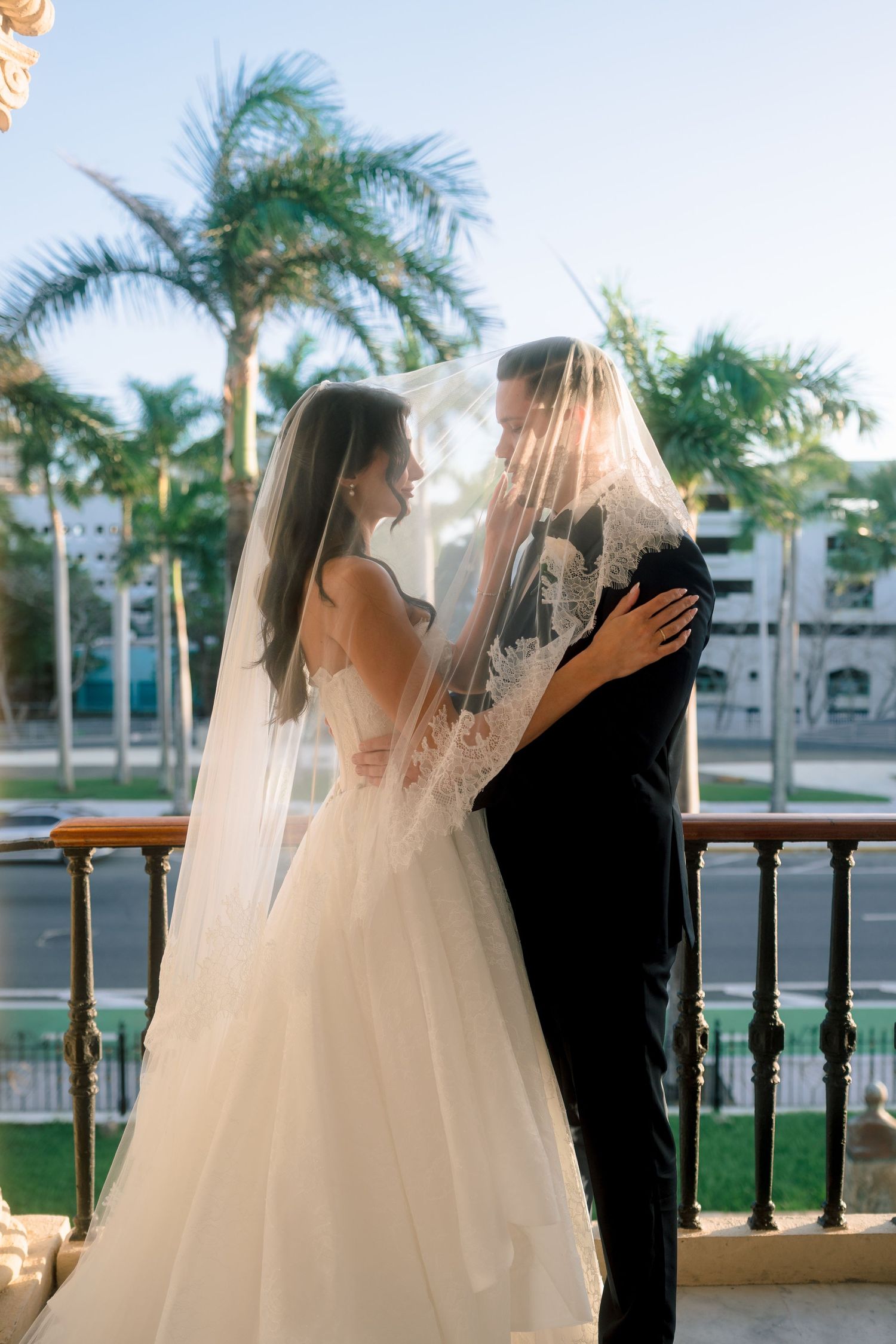Romantic couple shares intimate moment under bridal veil on sunlit balcony with palm trees in background.