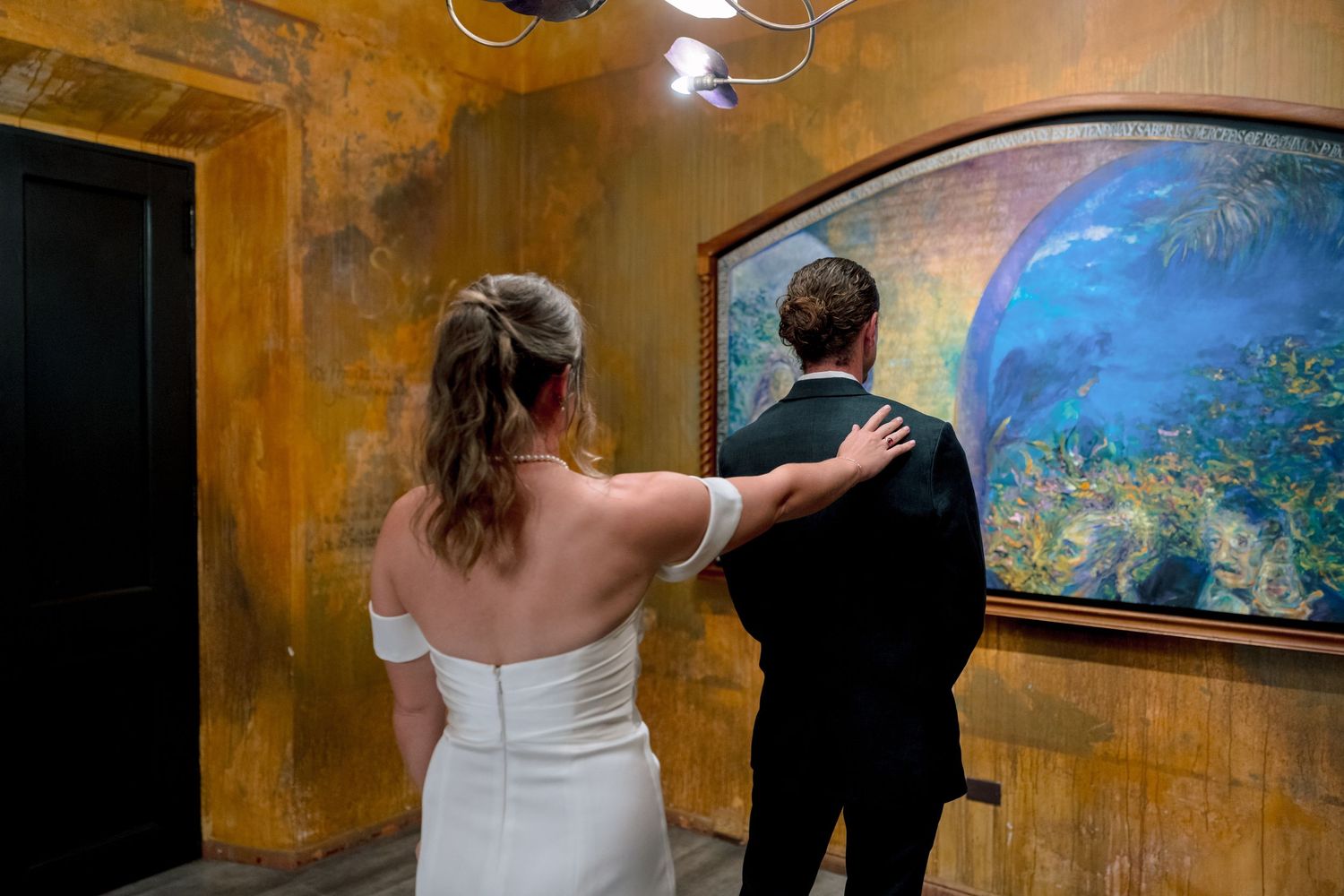Bride and groom in wedding attire viewing a colorful aquarium mural on a textured golden wall.