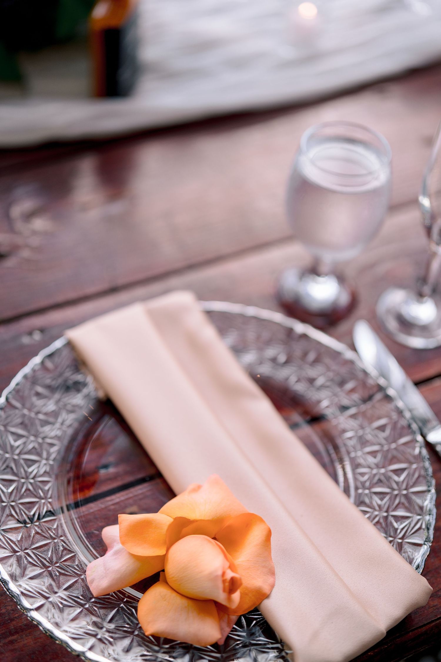Elegant crystal plate with peach rose petals and folded linen napkin on rustic wood table with glassware.