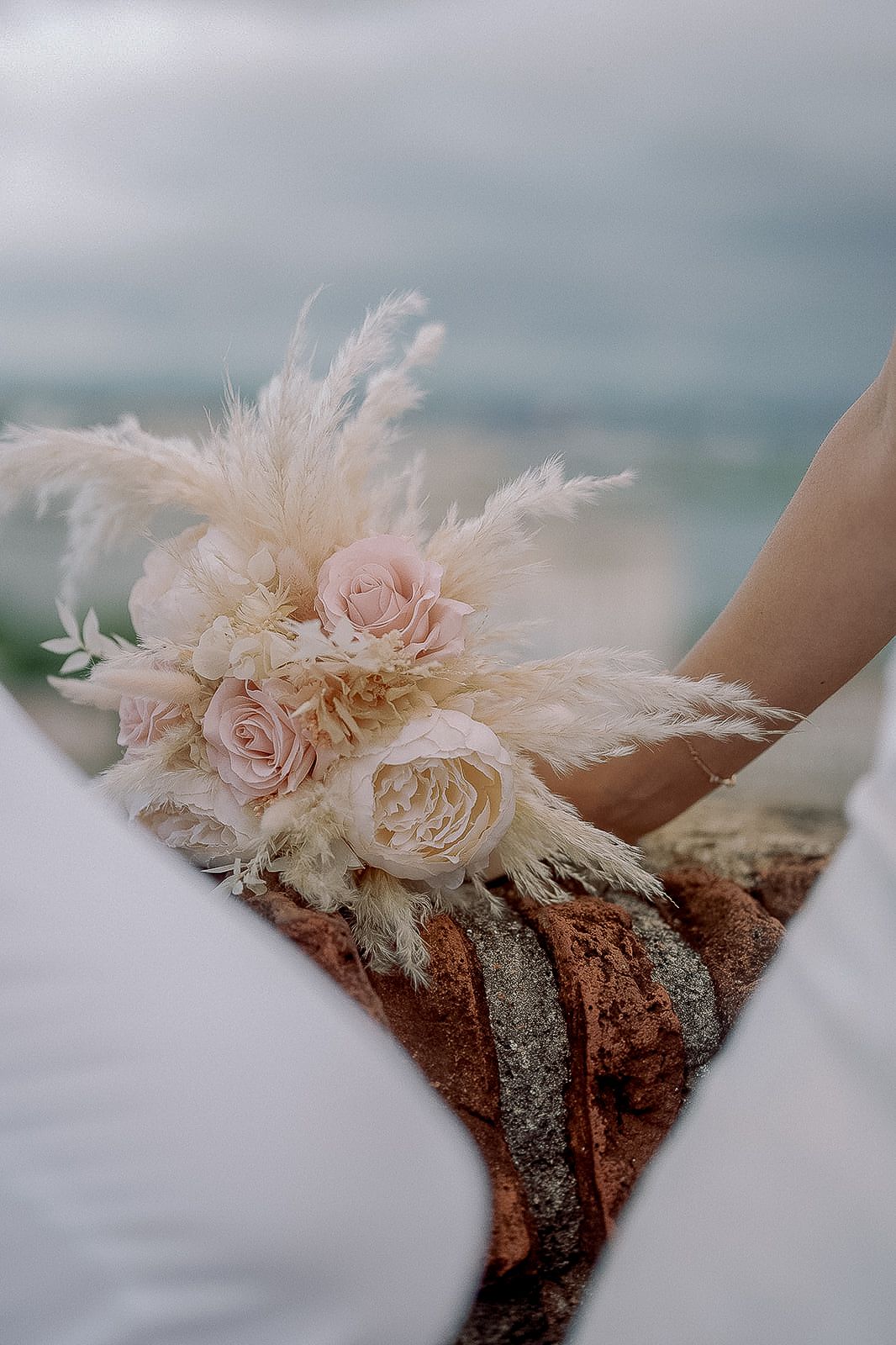 Bridal bouquet with blush pink roses and pampas grass held by bride near ocean backdrop.