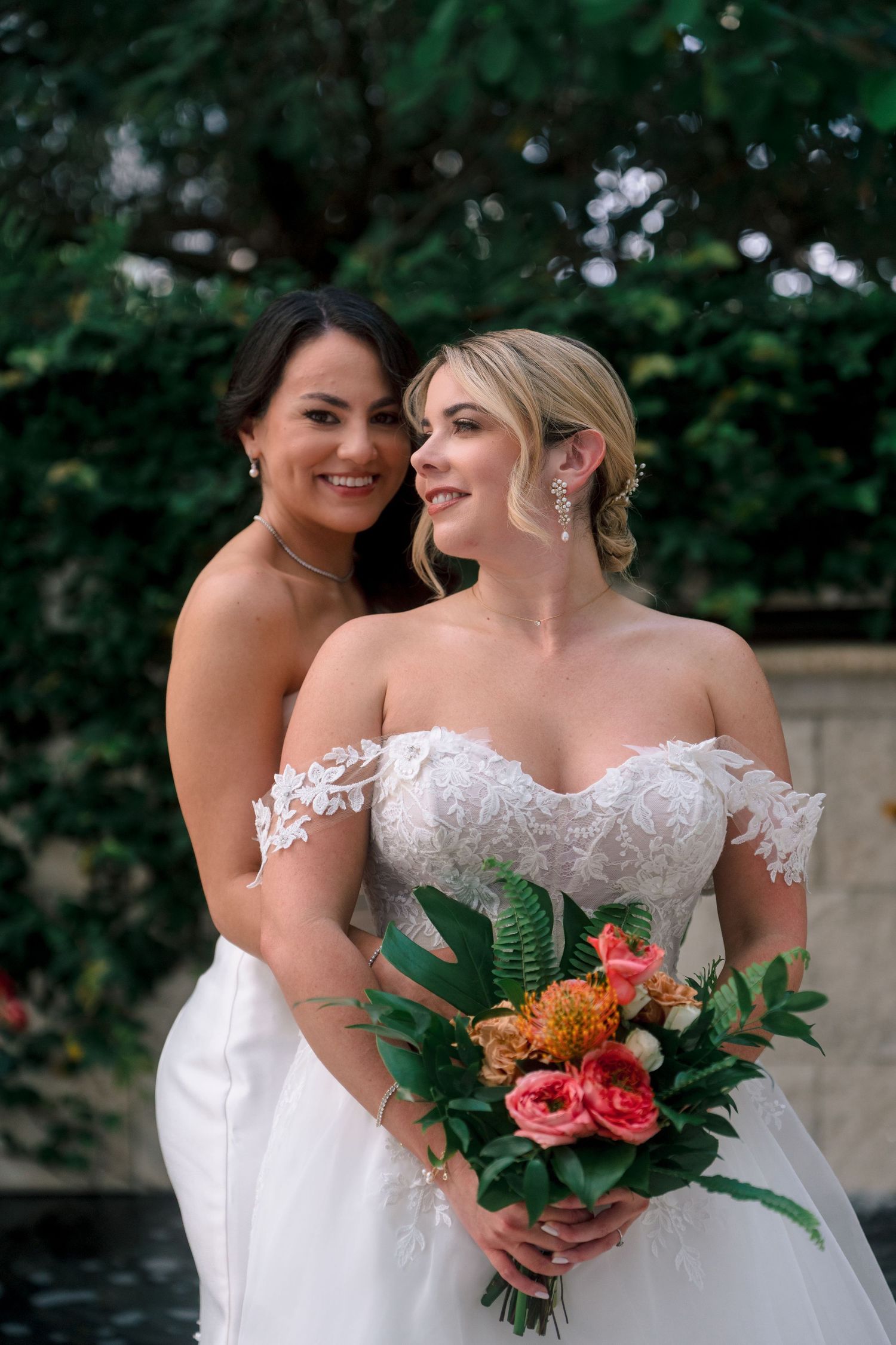 Two brides in white wedding gowns pose together, one holding a vibrant tropical bouquet with pink and orange flowers.