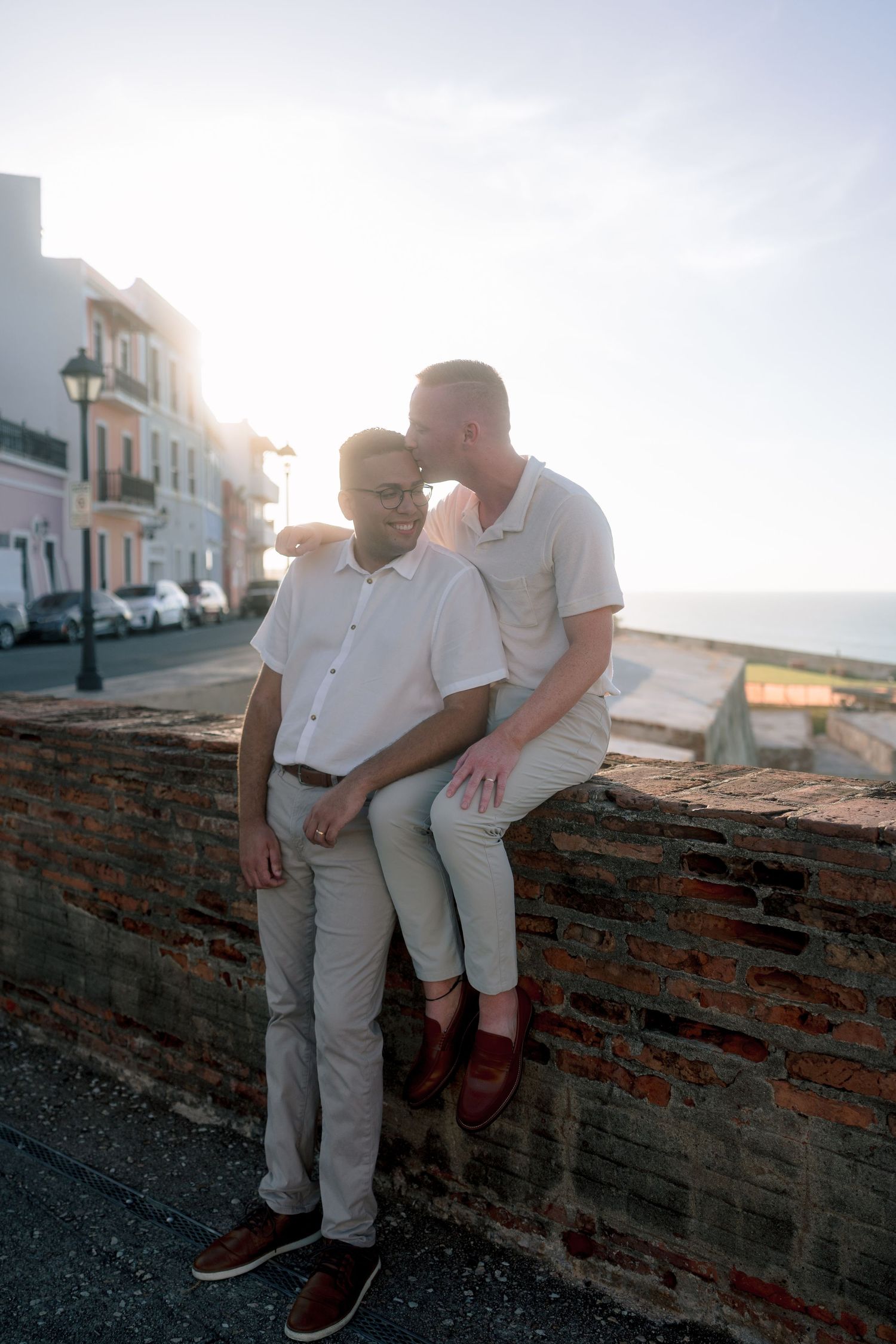 Two men share a tender moment on a brick wall in Old San Juan, Puerto Rico, with colorful buildings and ocean views.