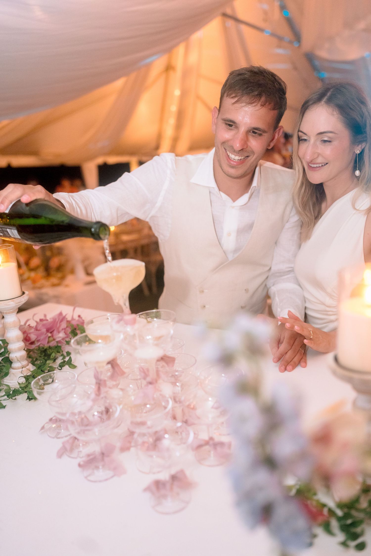 A smiling groom pours champagne into a tower of glasses beside his bride at a romantic wedding reception.