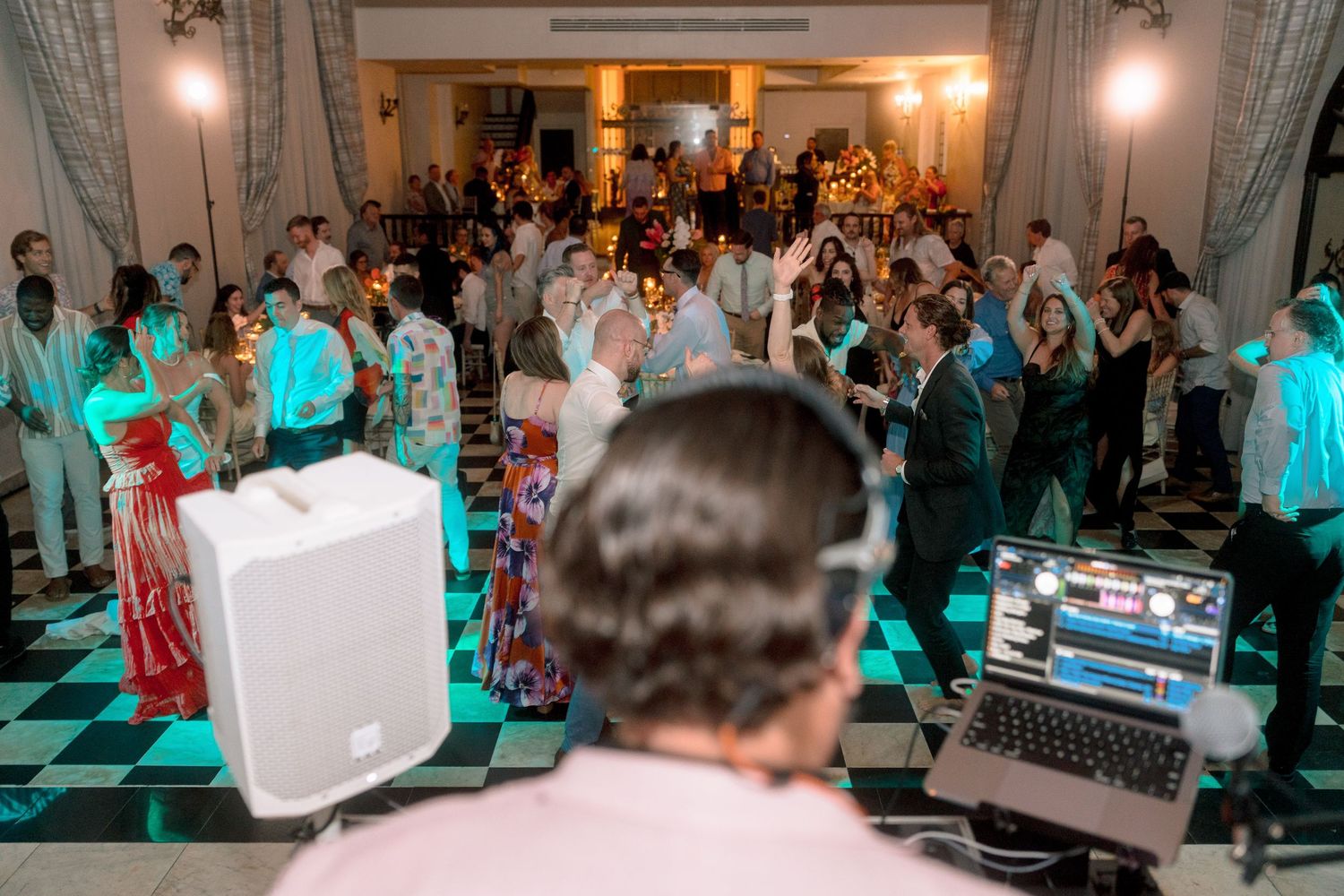 DJ performing at a lively wedding reception with guests dancing on a black and white checkered dance floor in an elegant venue.