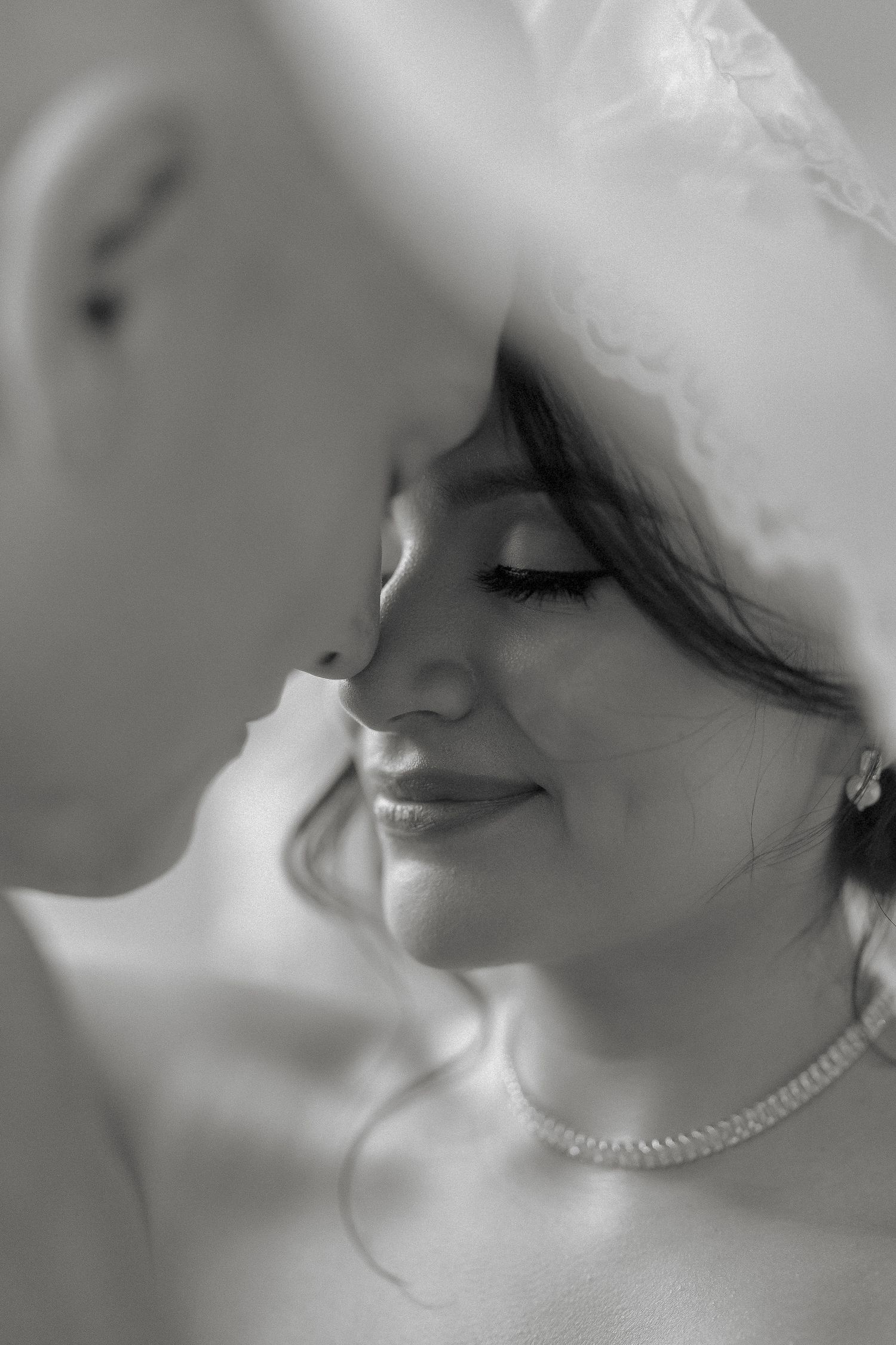 Black and white close-up of smiling bride wearing pearl necklace and lace veil with groom nearby.