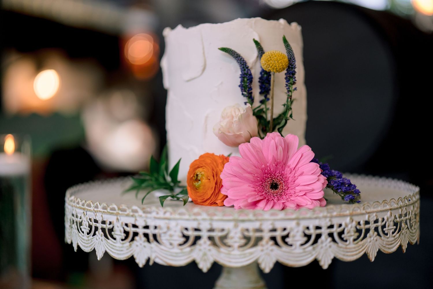 White wedding cake on ornate stand adorned with colorful fresh flowers including pink gerbera and orange ranunculus.