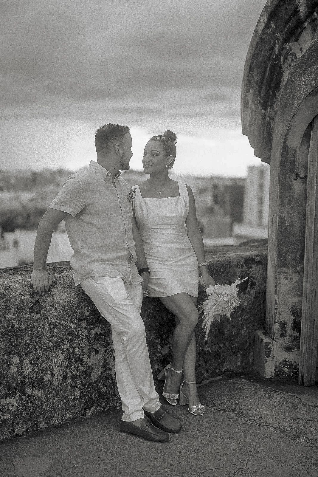 Black and white photo of a couple in white attire posing romantically on a historic stone fortress wall.