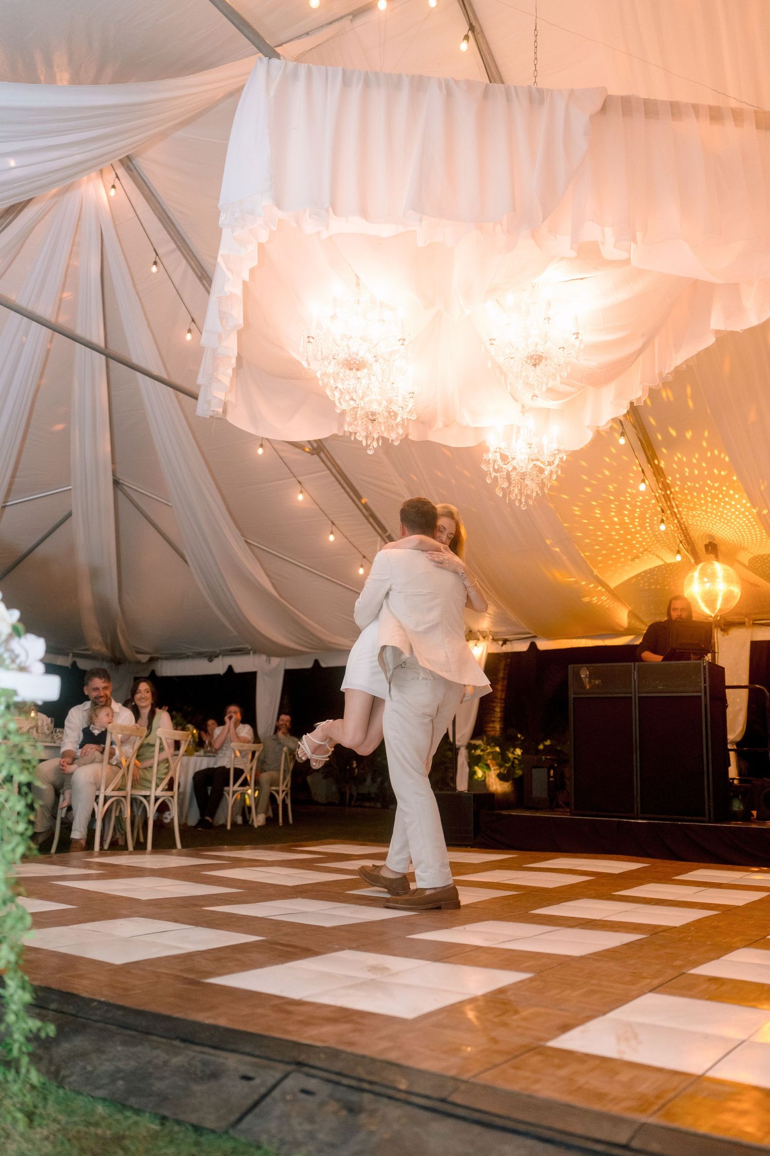 Couple shares first dance under elegant white tent with chandeliers and string lights at outdoor wedding reception.