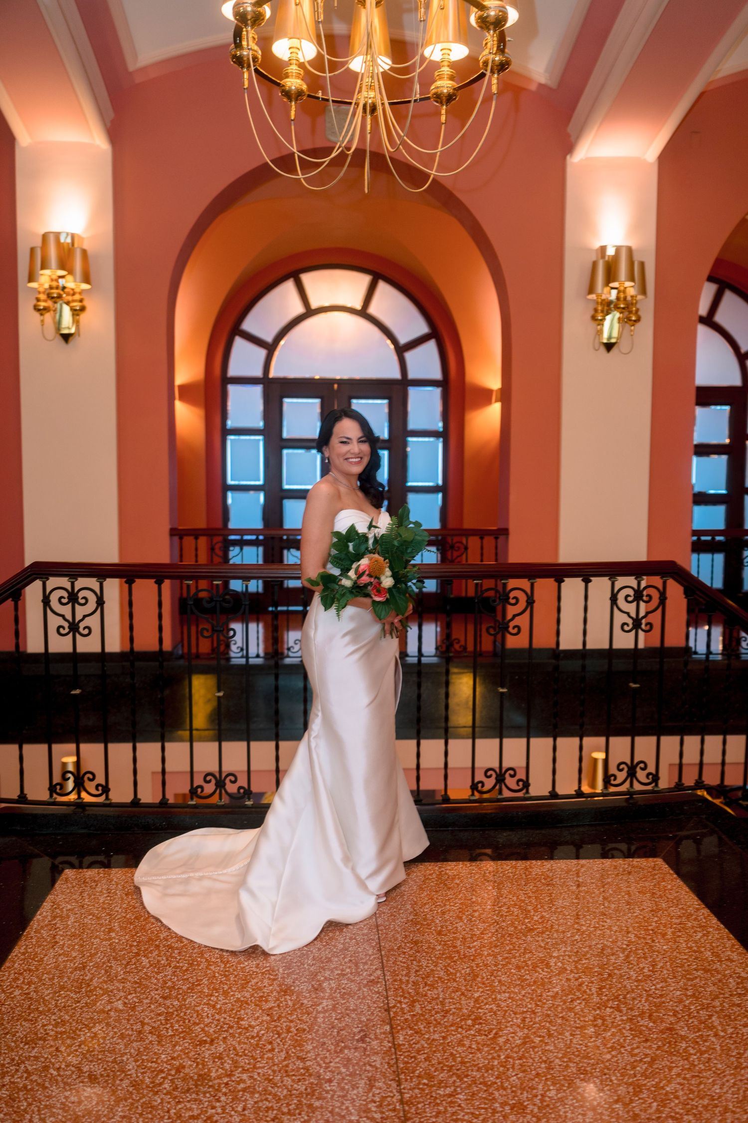 Bride in white satin gown holding bouquet stands on marble floor in elegant venue with arched windows and chandelier.