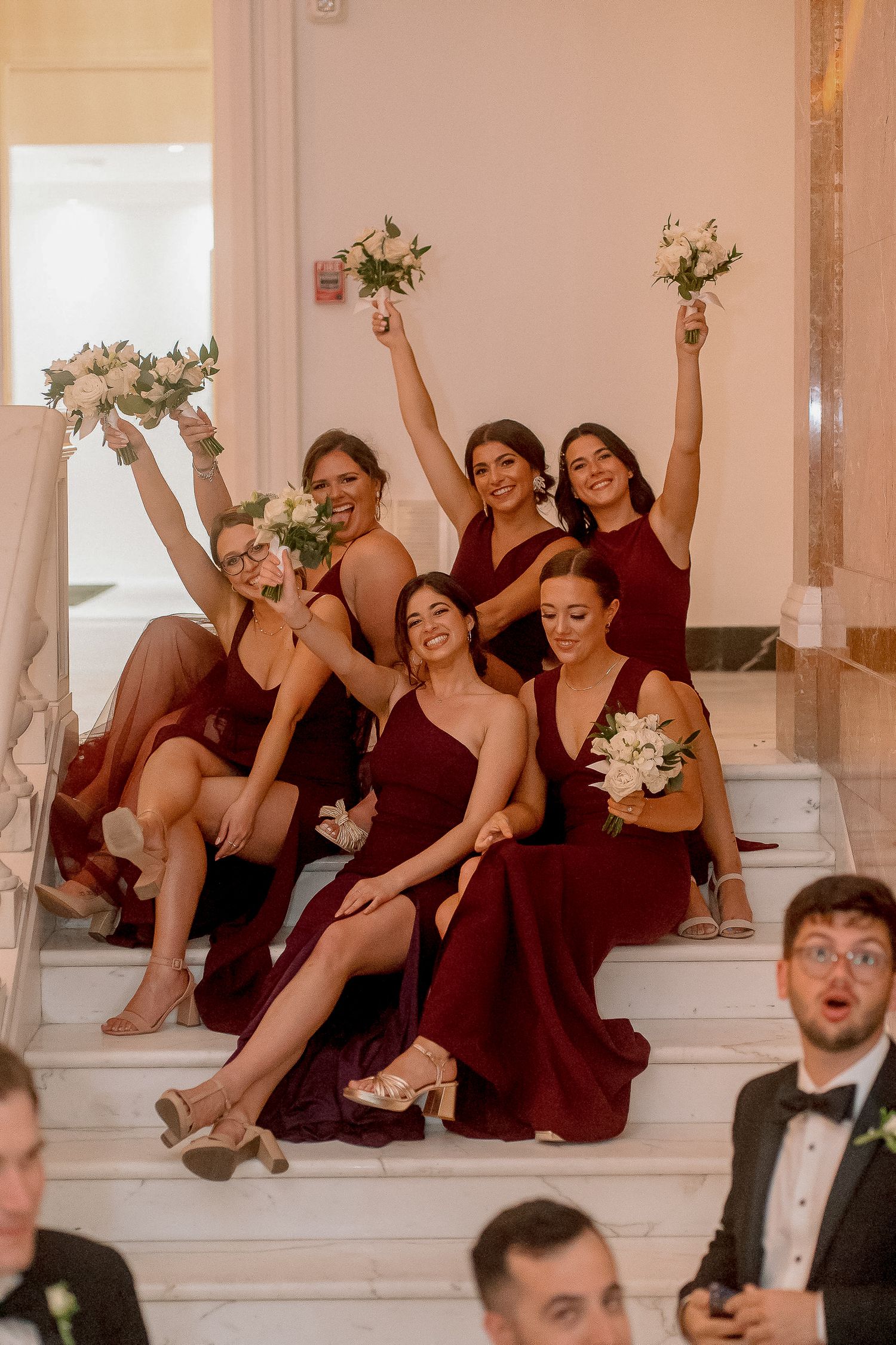 Six bridesmaids in burgundy gowns pose on marble stairs raising white bouquets in celebration at a wedding.