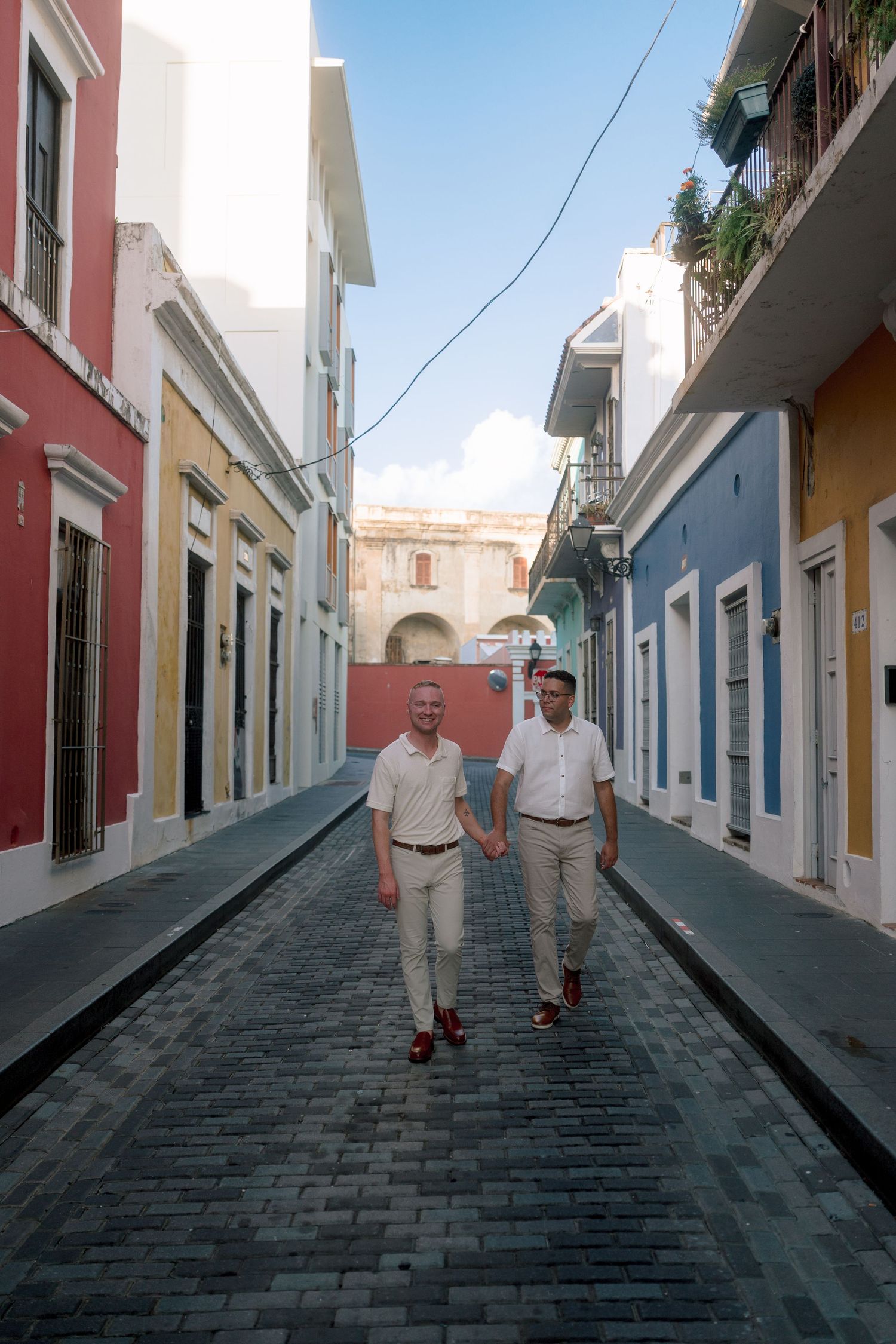 Two men in matching white shirts walk hand-in-hand down a colorful cobblestone street in Old San Juan.