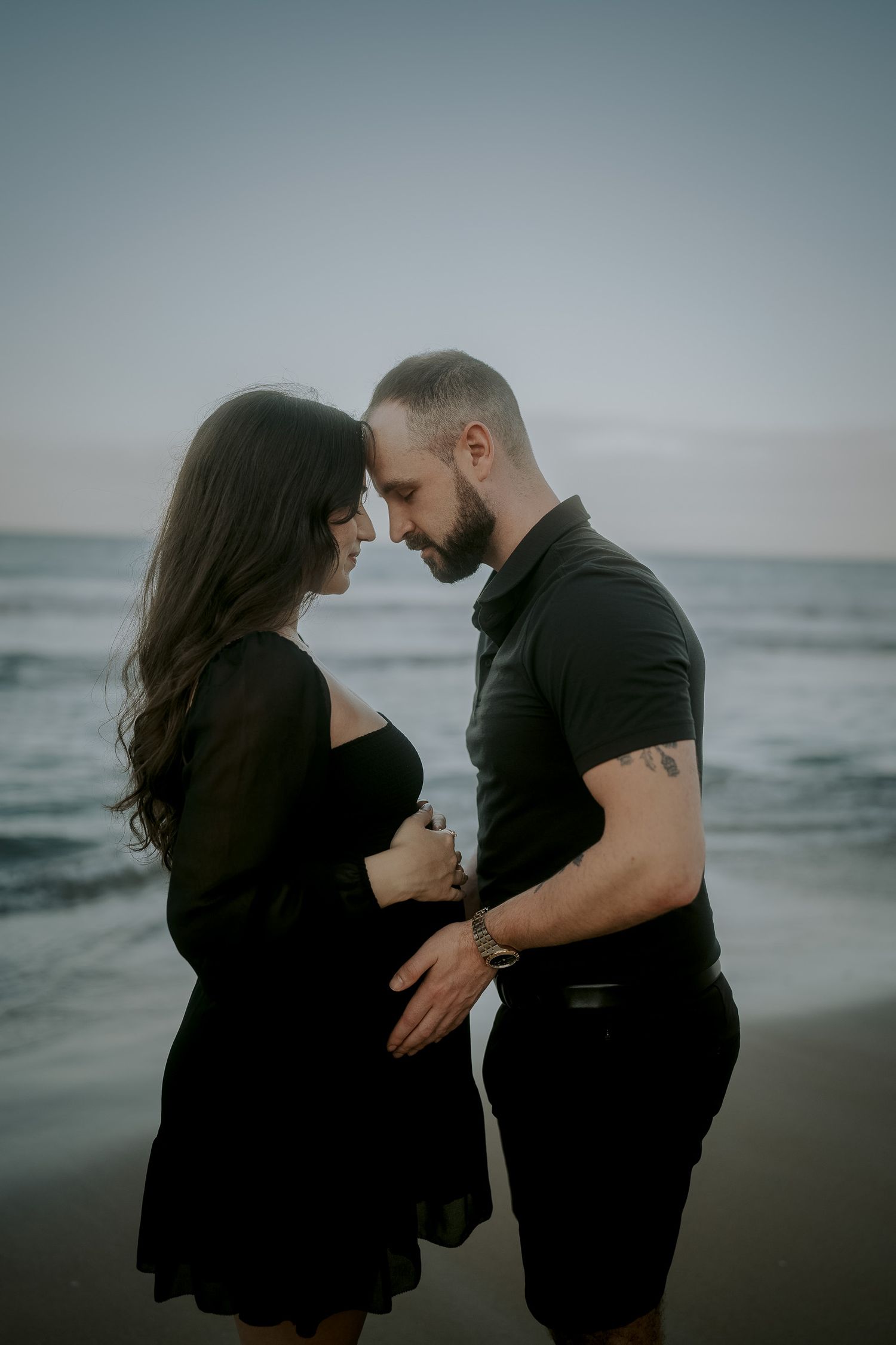 Couple in black outfits sharing tender moment on beach at dusk, man gently touching pregnant woman's belly.
