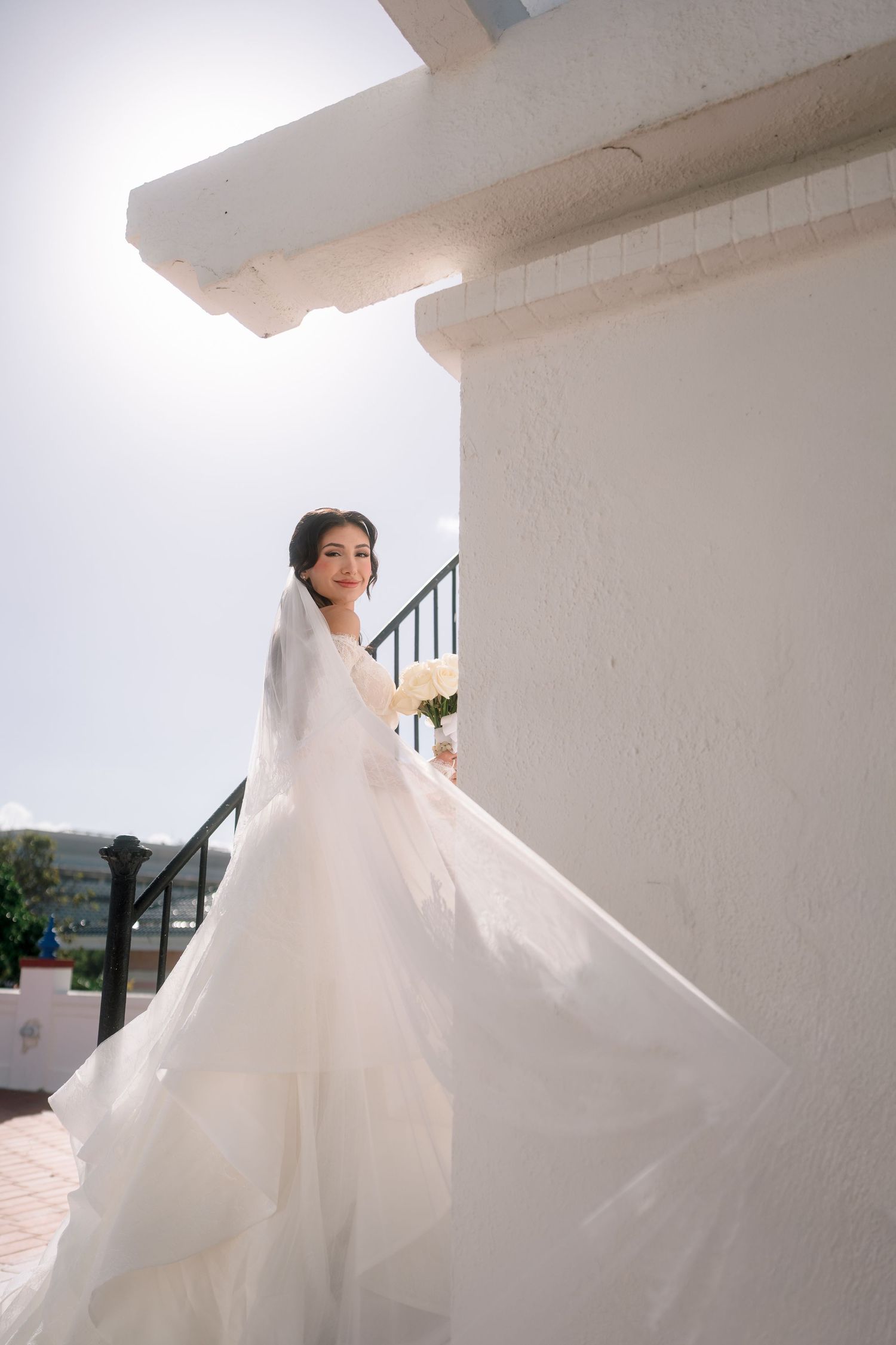 Bride in flowing white wedding gown with veil poses gracefully outdoors against white wall in bright sunlight.