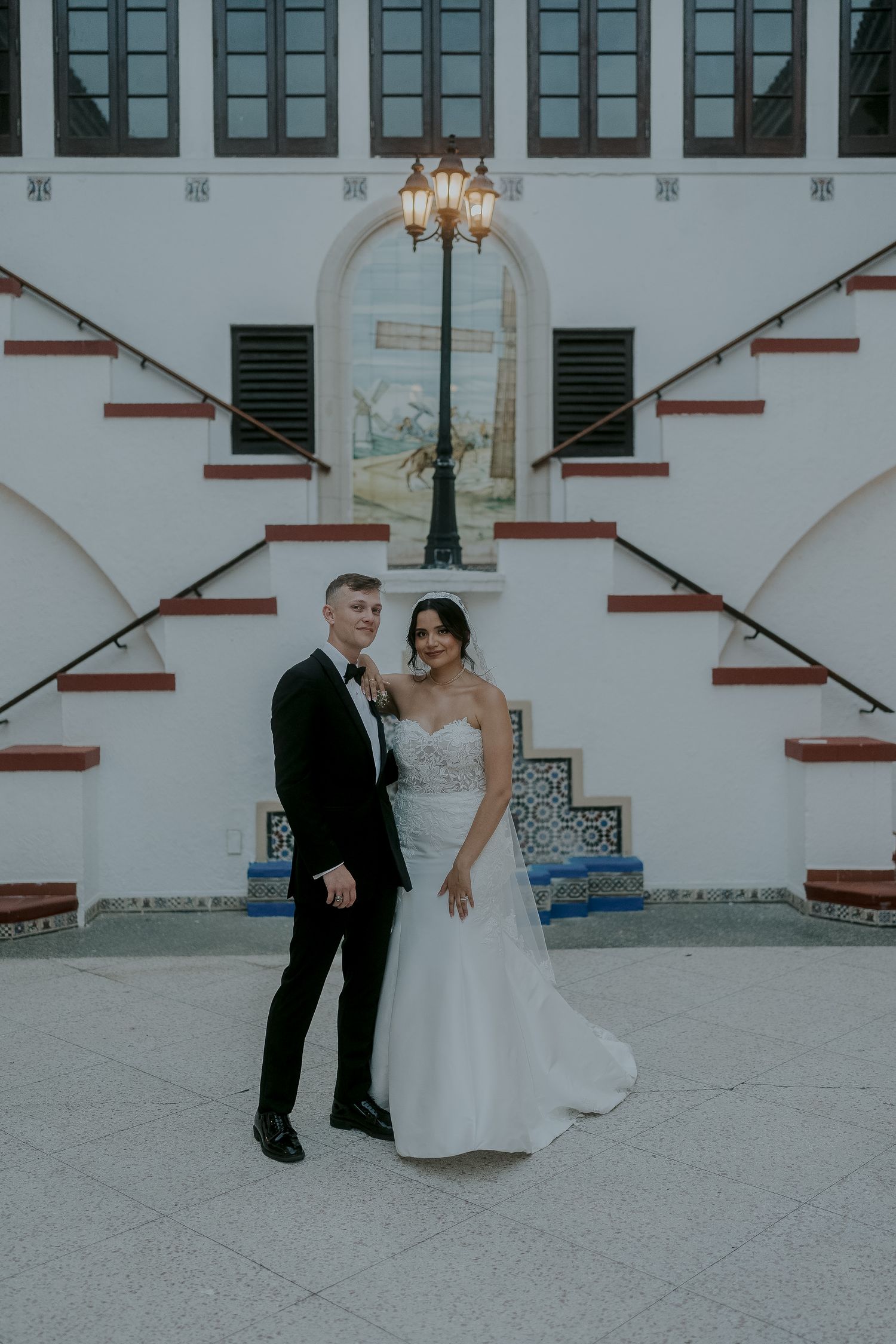 Bride and groom posing together in front of white Spanish colonial building with decorative staircase.