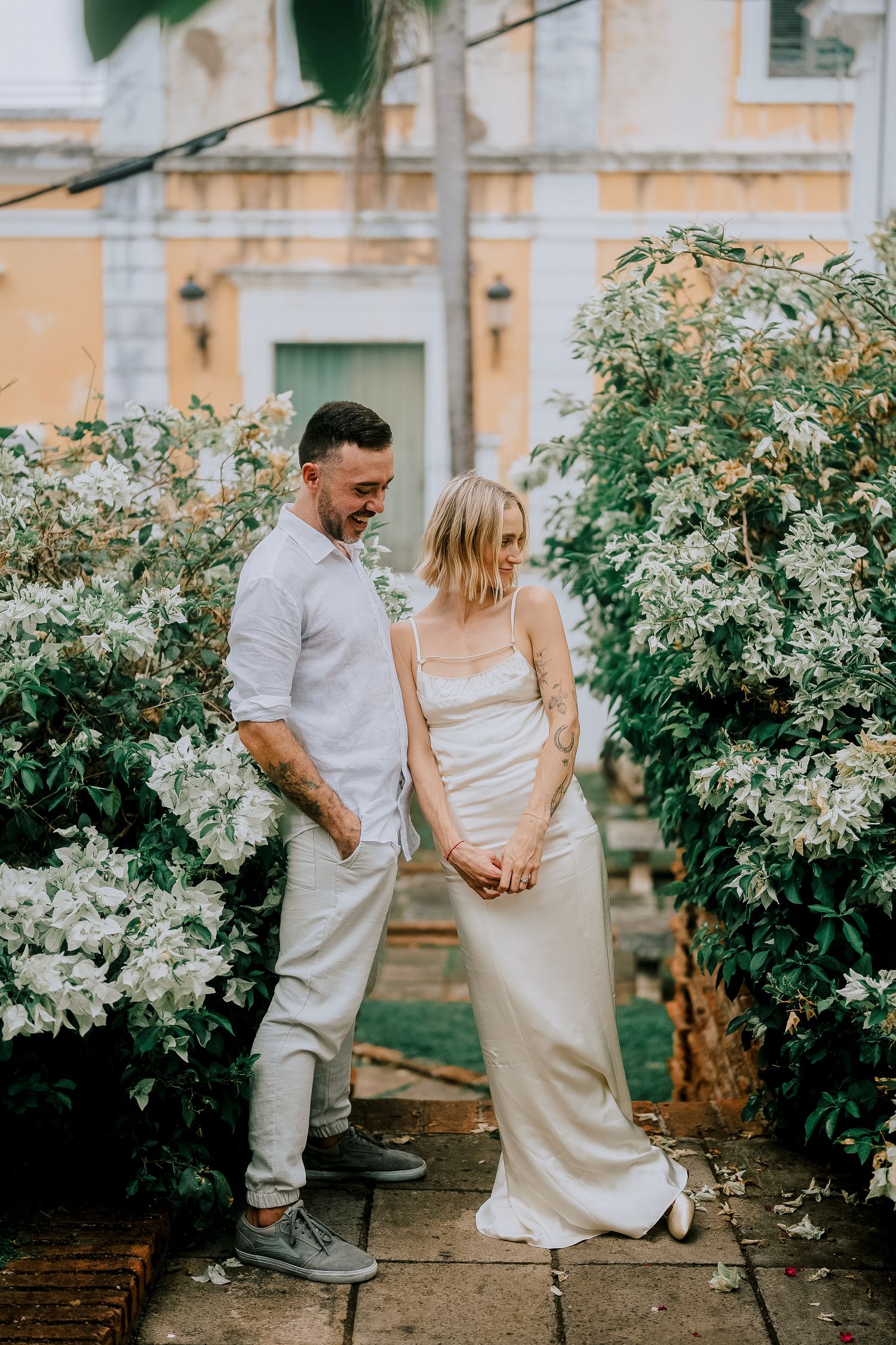Couple in wedding attire holding hands among white flowering bushes near a yellow building.