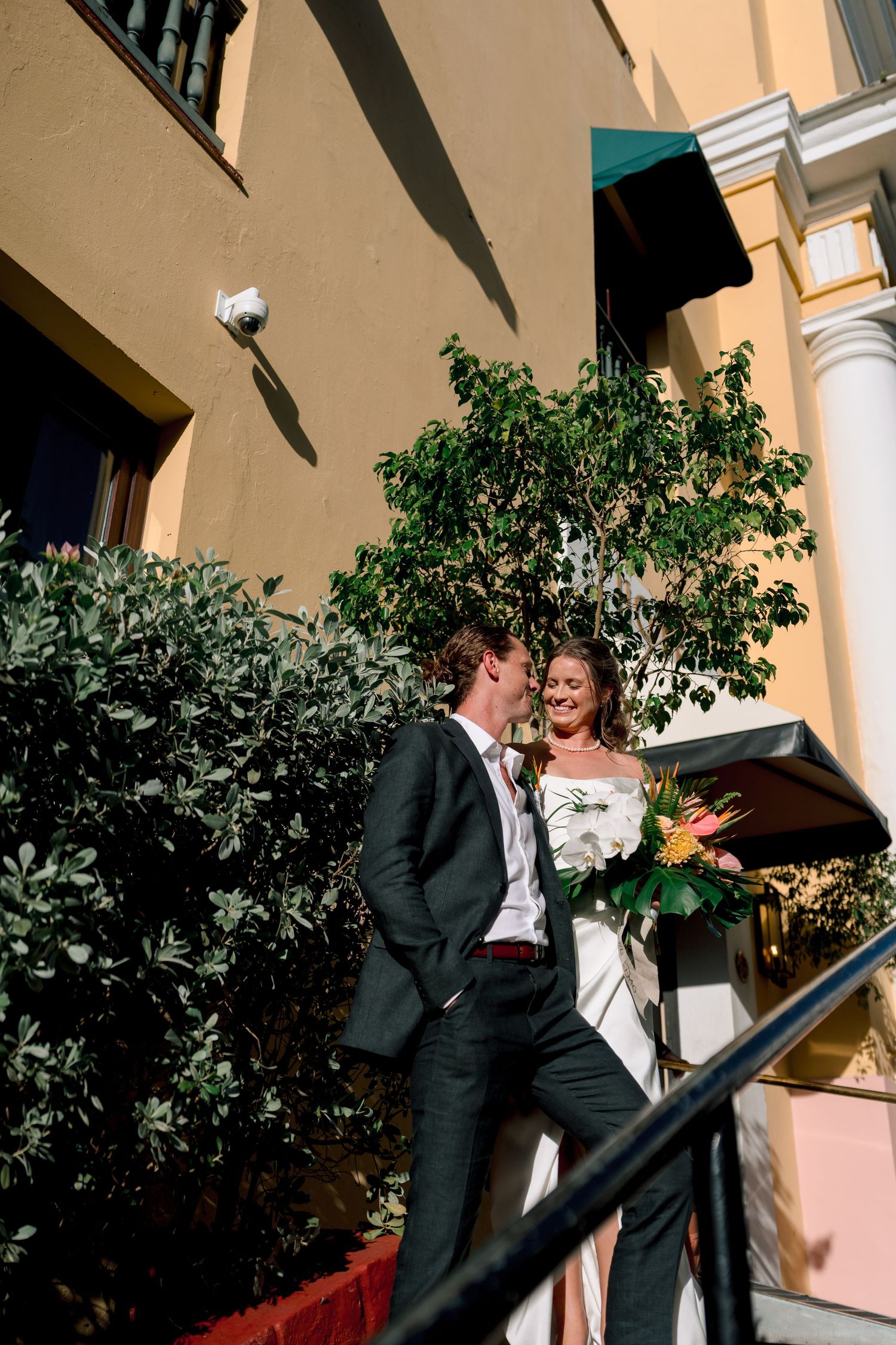 A smiling bride in a white gown holds a tropical bouquet beside a groom in a dark suit on sunlit outdoor steps.
