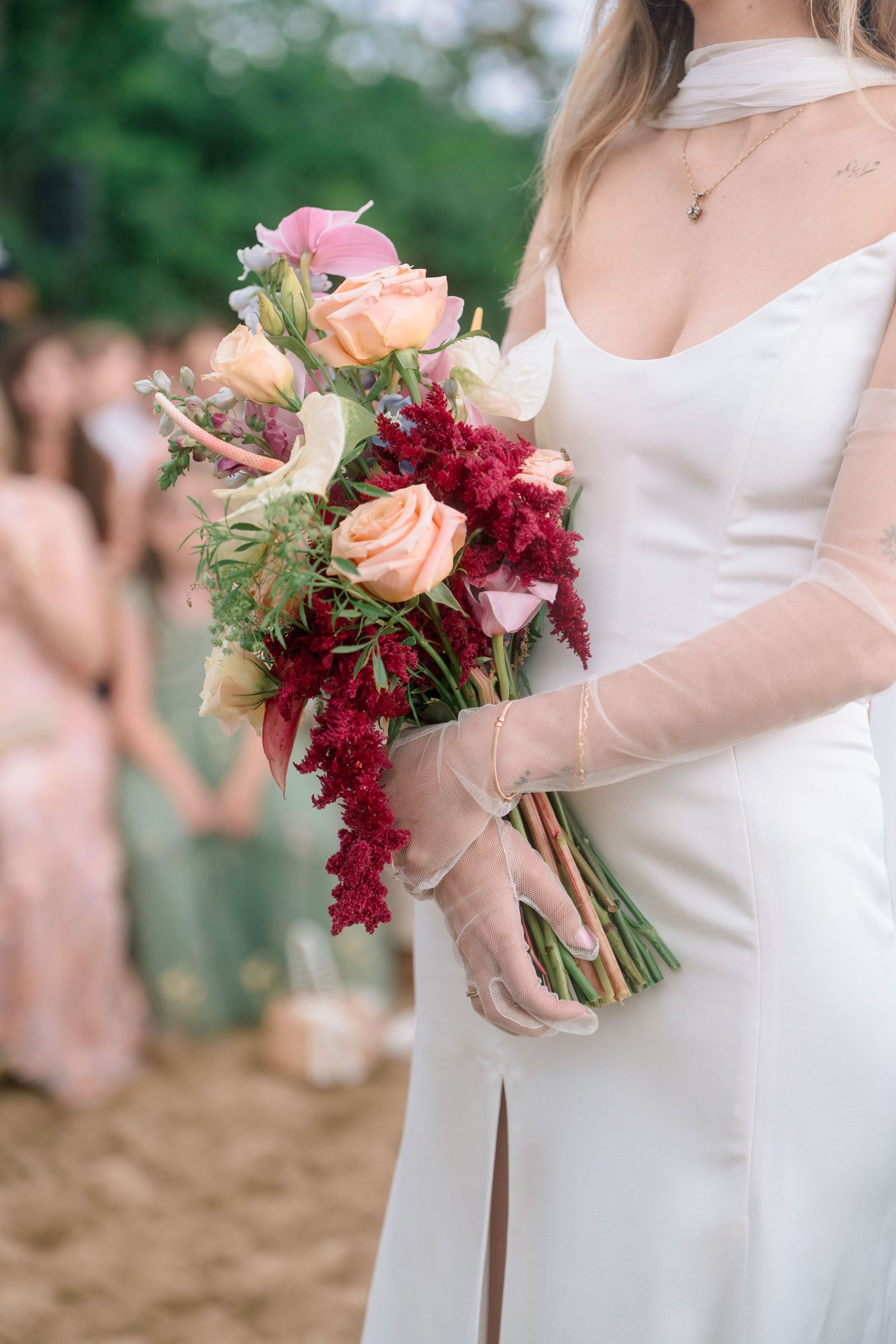 Bride in white gown with sheer gloves holding vibrant red and peach floral bouquet at outdoor wedding.