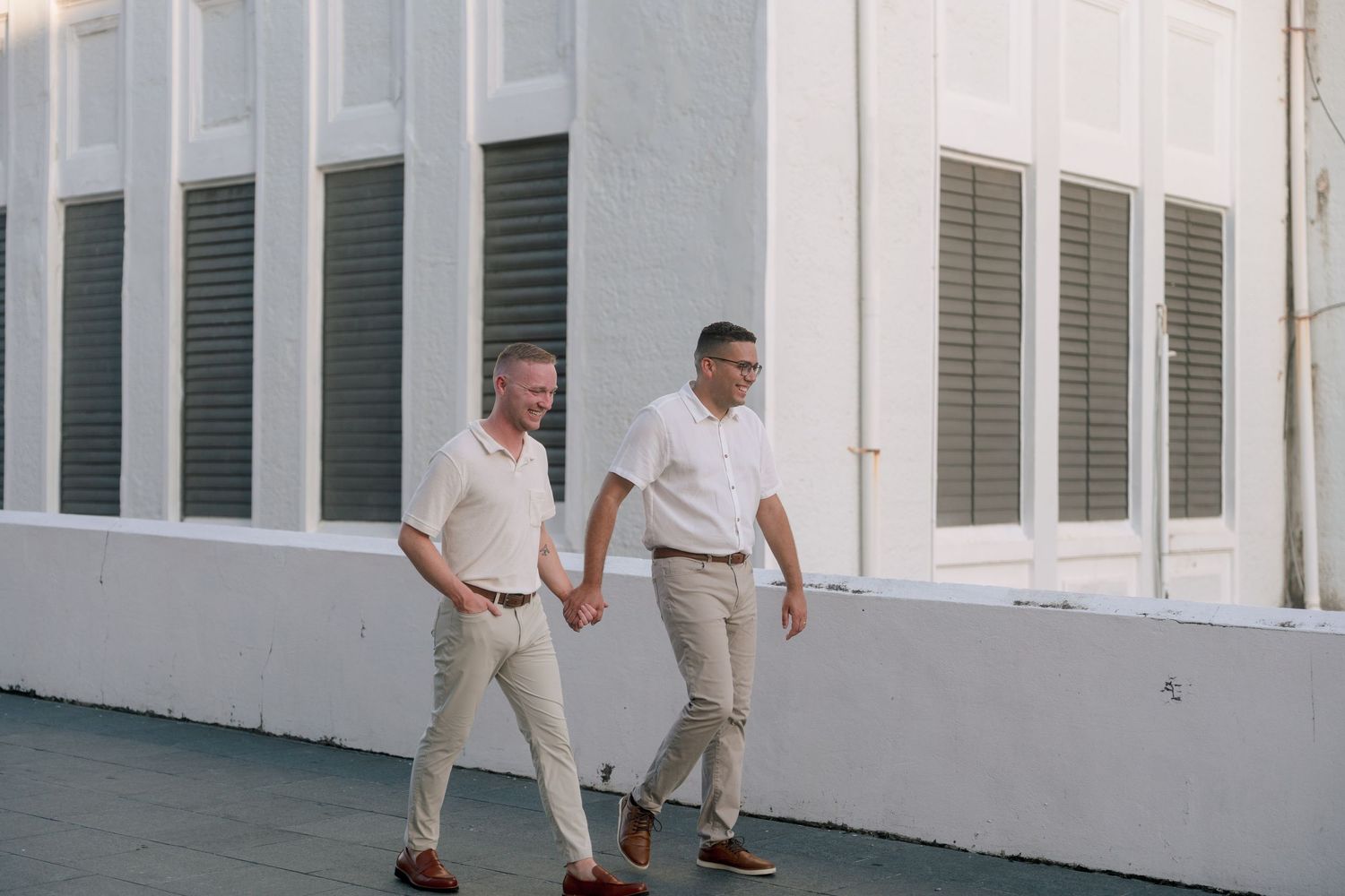 Two men in white shirts and beige pants walk hand in hand outside a white building with shuttered windows.