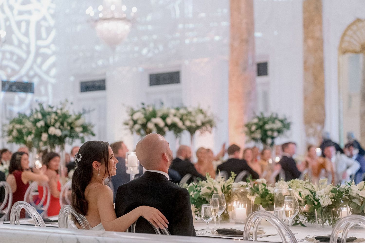 Bride and groom seated at sweetheart table during elegant white wedding reception.