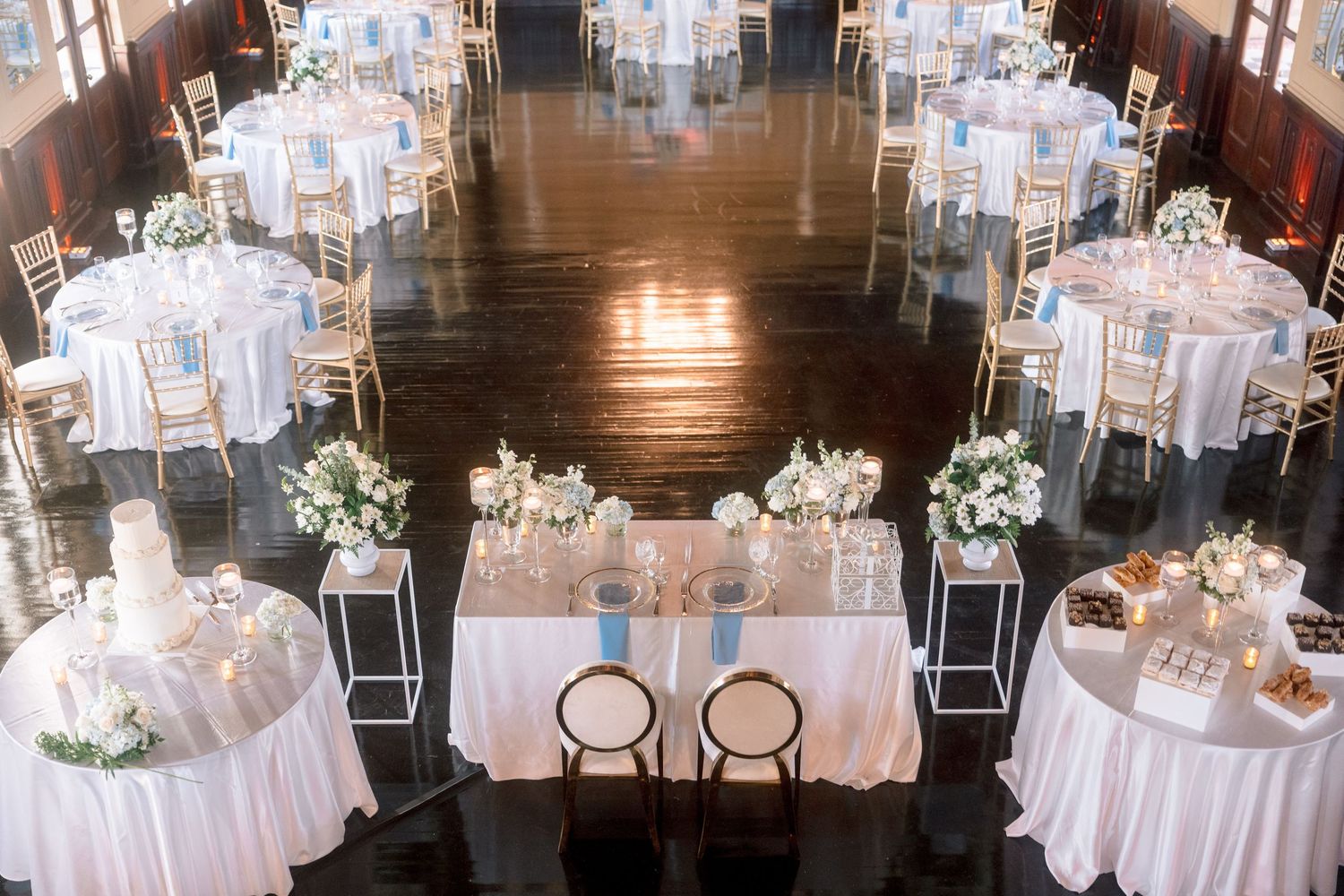 Elegant wedding reception with round tables, white linens, floral centerpieces, and a sweetheart table on dark hardwood floors.