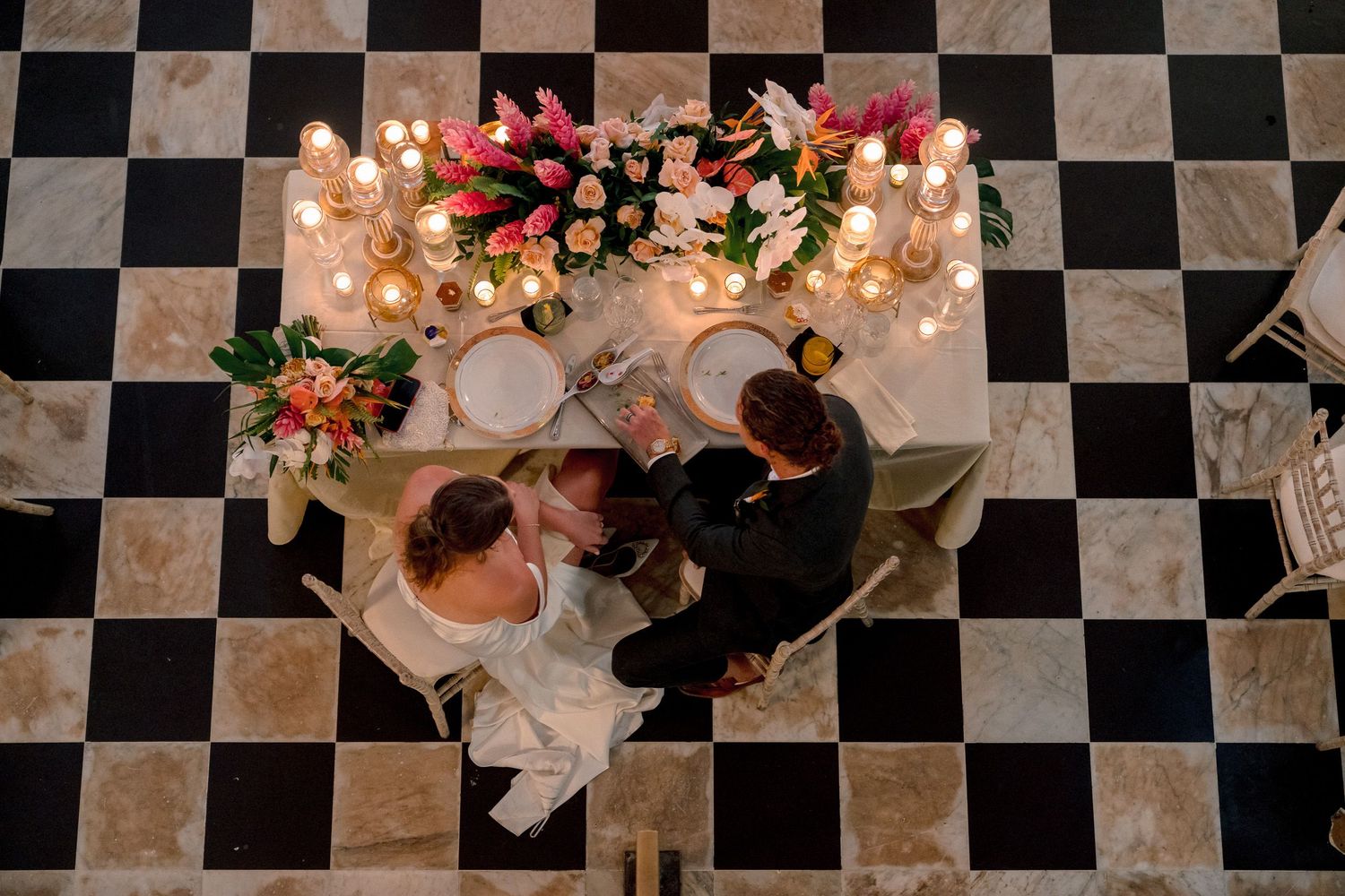 Aerial view of bride and groom at candlelit sweetheart table with floral centerpiece on checkered floor.