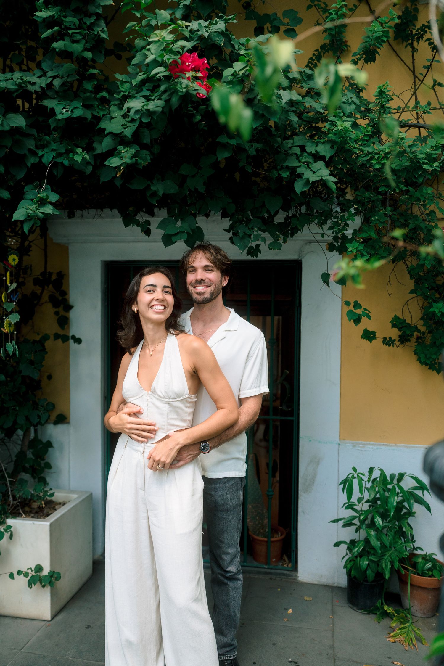 Smiling couple embracing in white outfits under lush green vines at a Mediterranean villa entrance.