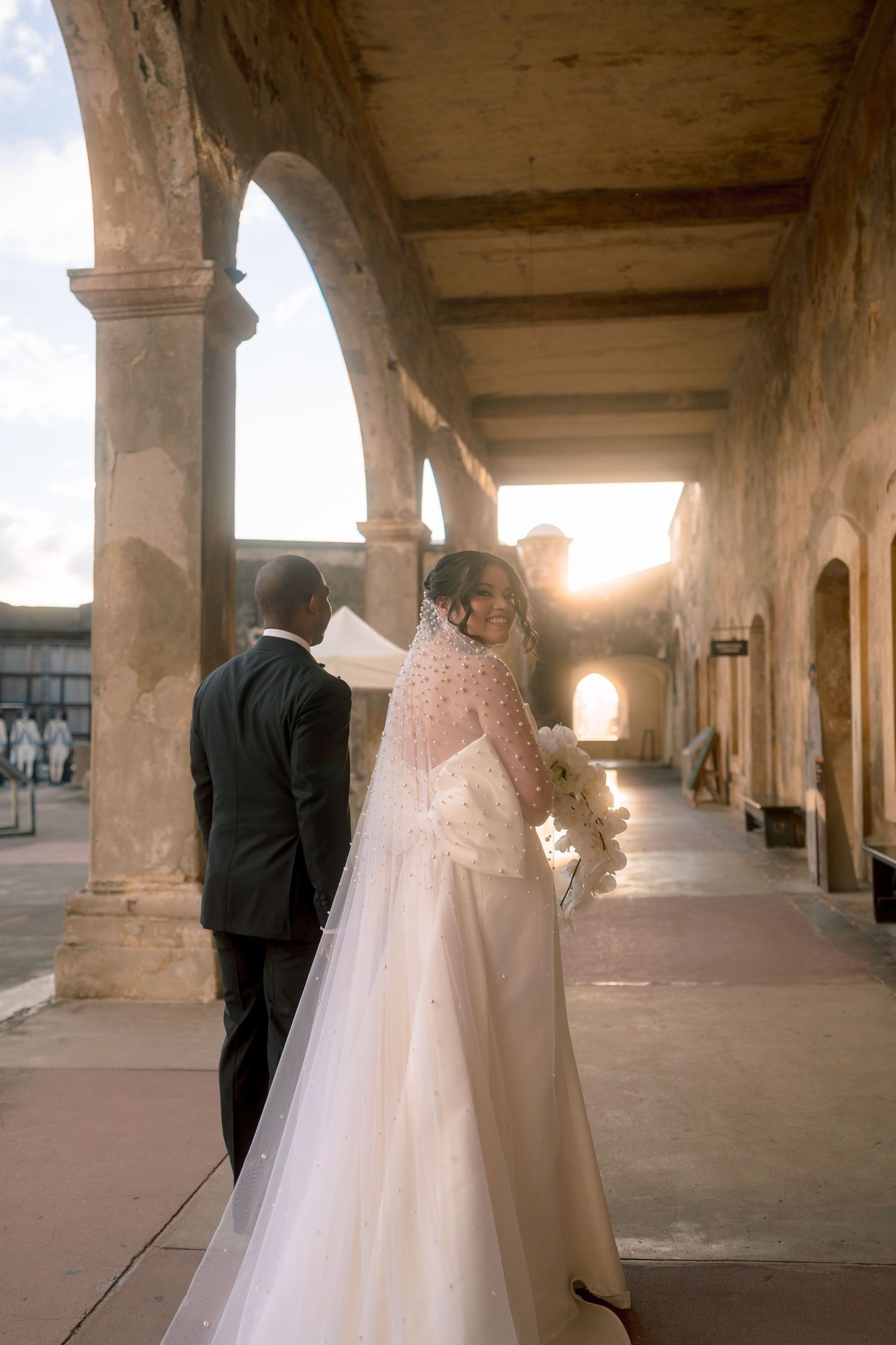 Bride in pearl-adorned veil and gown walks with groom in tuxedo through sunlit stone archway corridor.