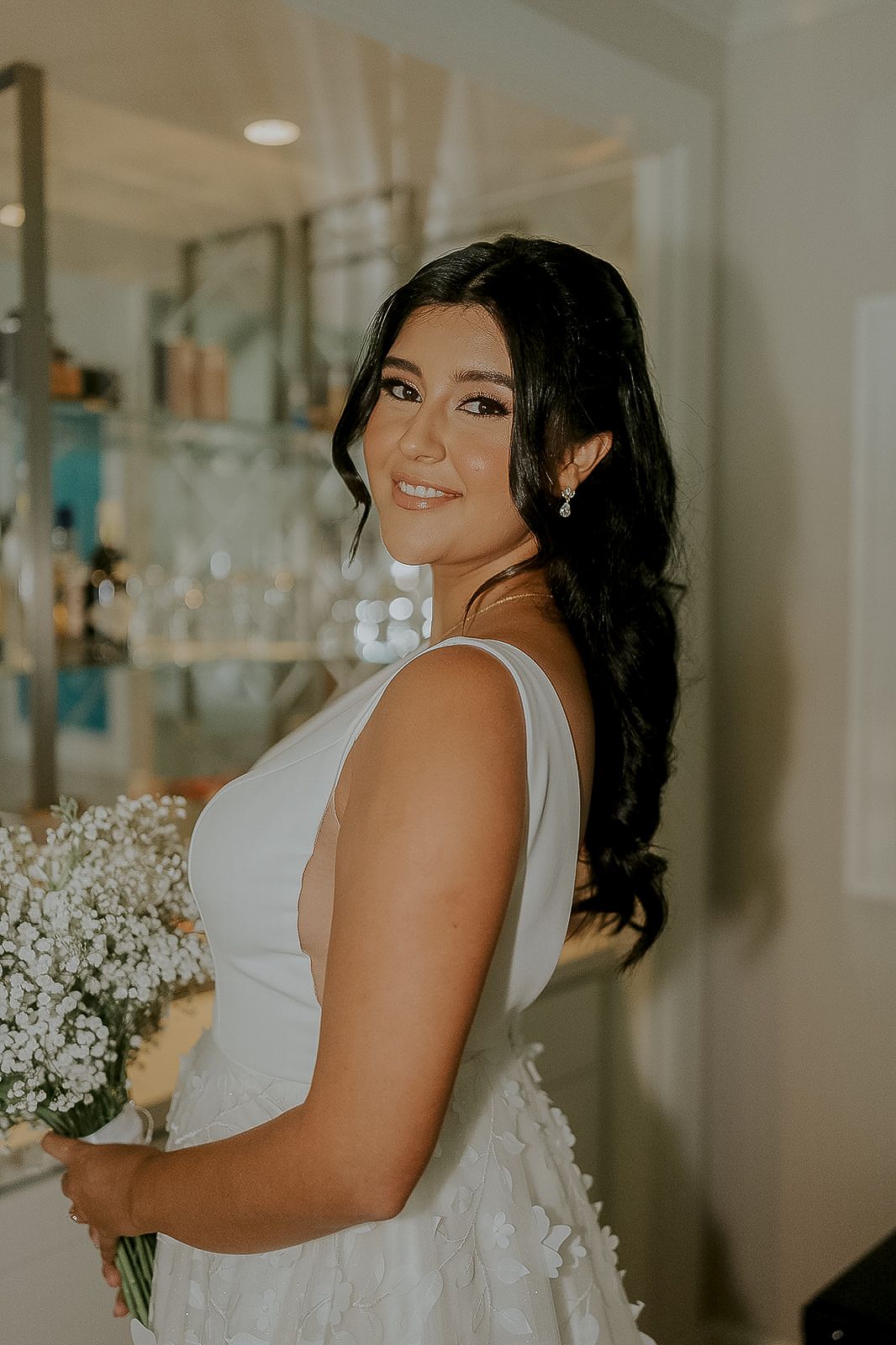 Bride in white gown holding baby's breath bouquet, smiling over shoulder with elegant dark wavy hair.