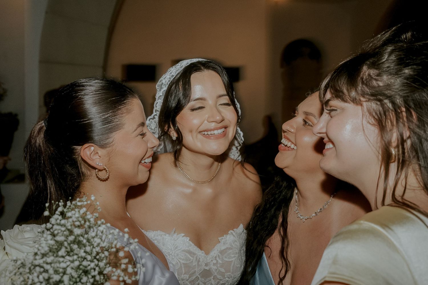 Bride in lace gown laughing joyfully with three bridesmaids in an intimate group hug at a wedding celebration.