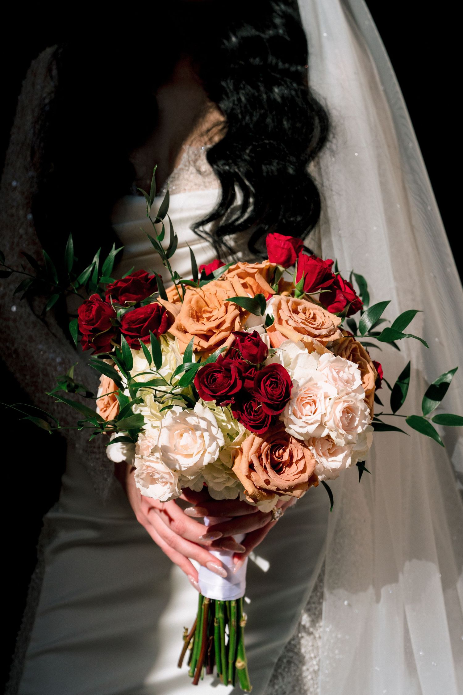 Bride holding lush bouquet of red, peach, and white roses with greenery accents.
