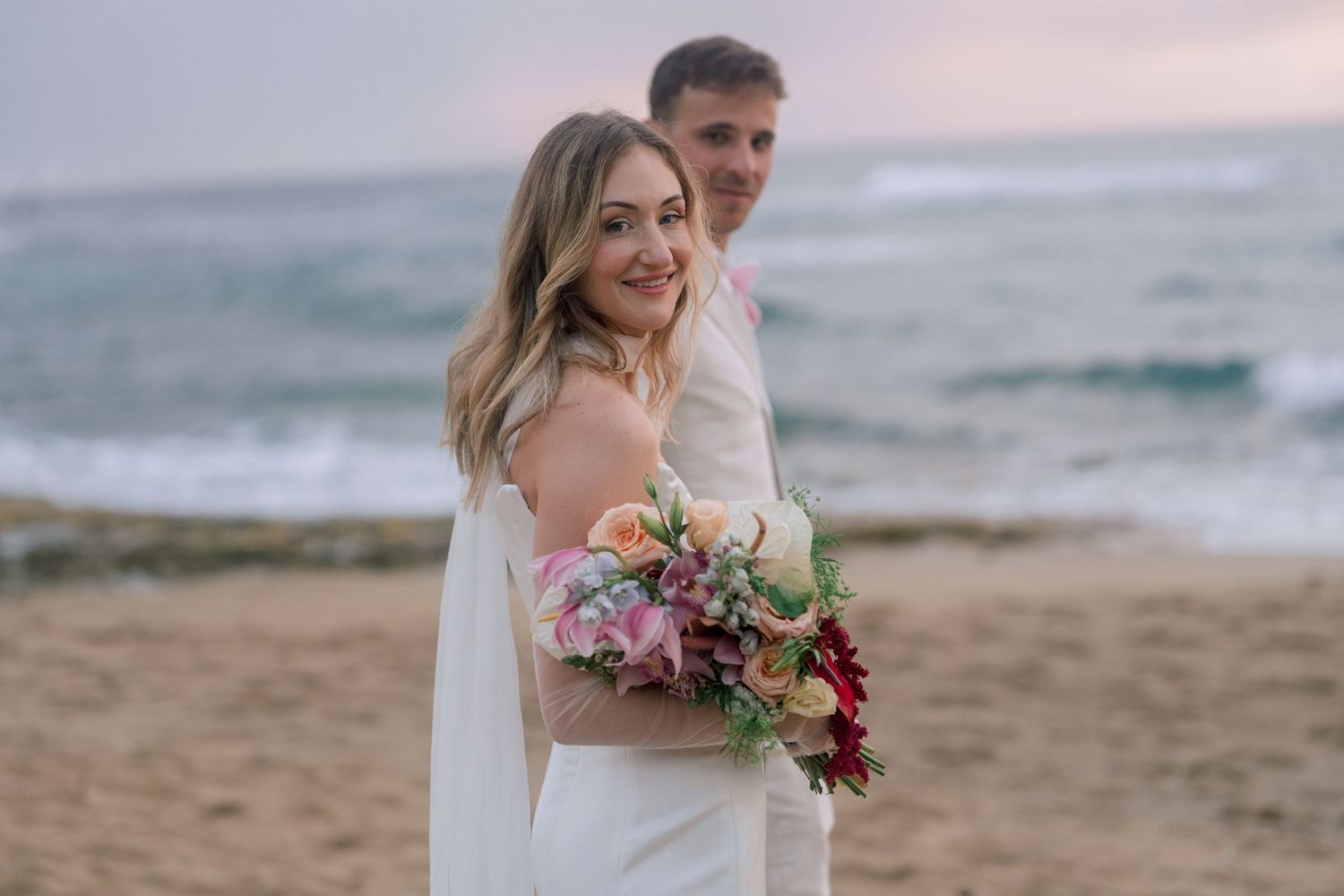 Bride and groom pose on a sandy beach at sunset, bride holding a colorful bouquet with ocean waves in the background.