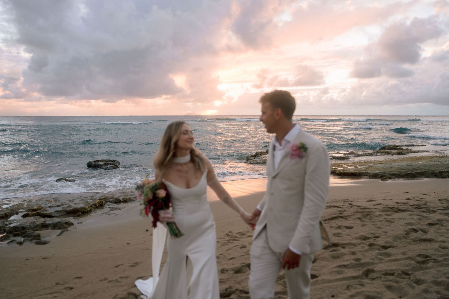 Bride and groom holding hands on a sandy beach at sunset, waves and colorful sky in the background.