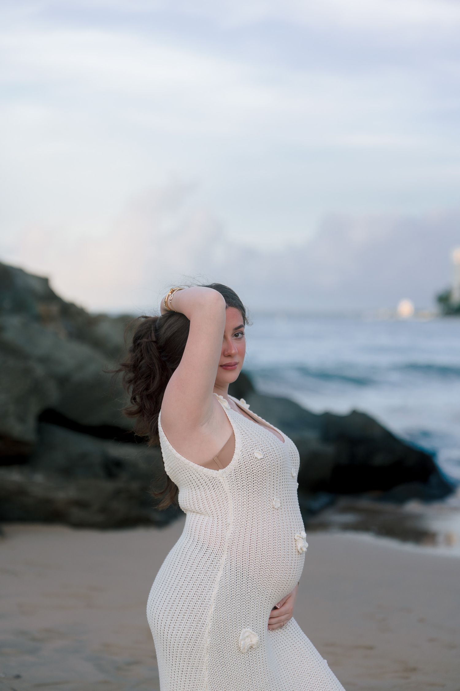 Pregnant woman in white knit dress poses gracefully on a sandy beach with rocky shores and ocean waves at dusk.