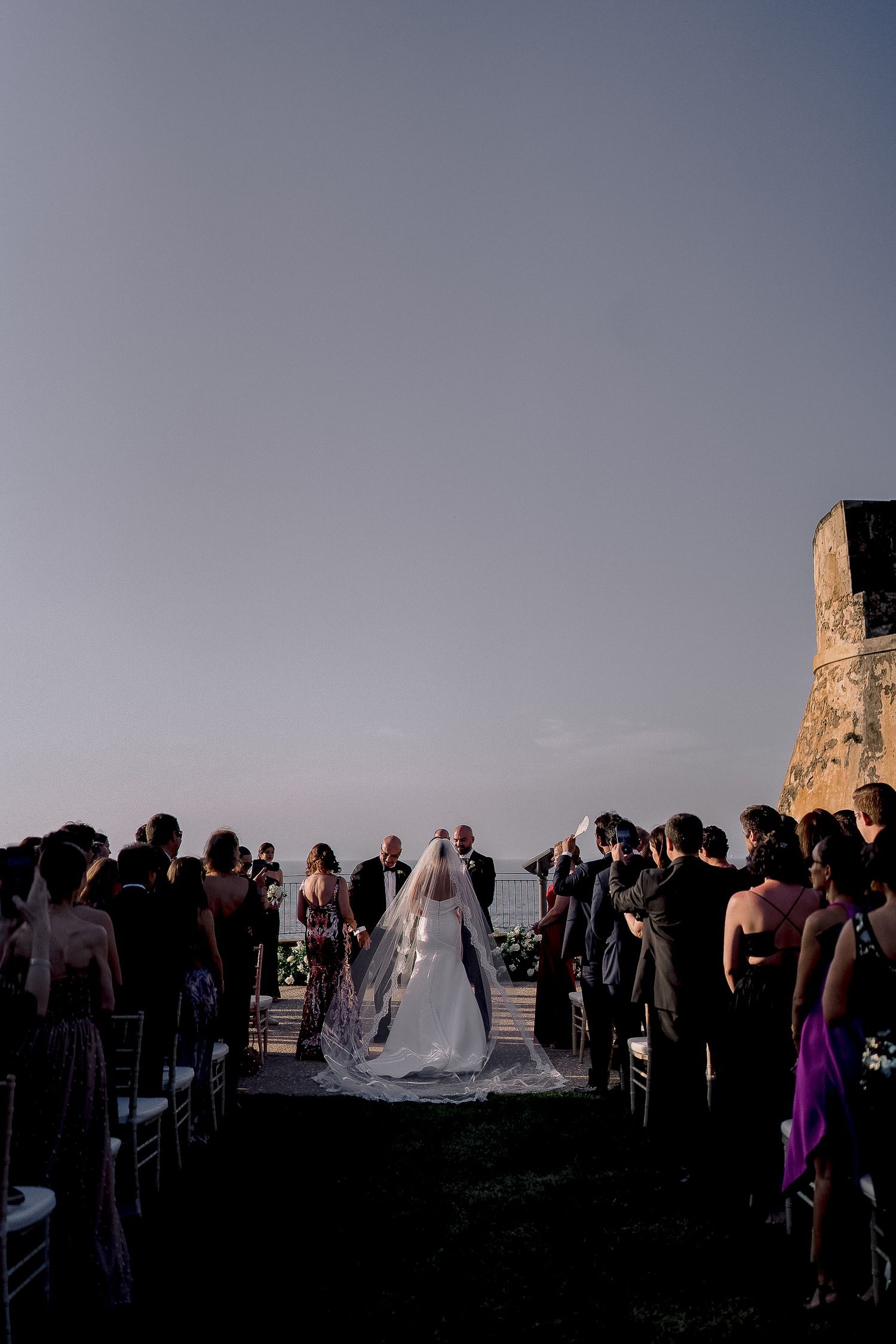 Bride walking down outdoor aisle at sunset wedding ceremony with guests seated on both sides near stone wall.