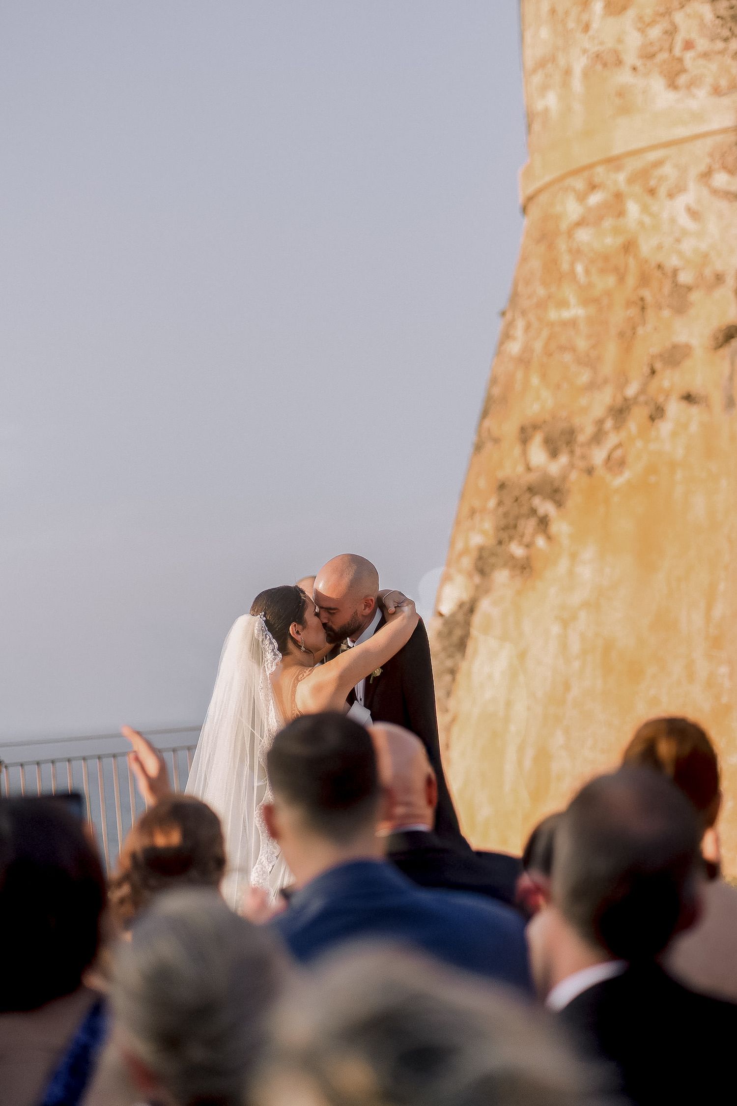 Bride and groom share first kiss surrounded by wedding guests at outdoor ceremony.