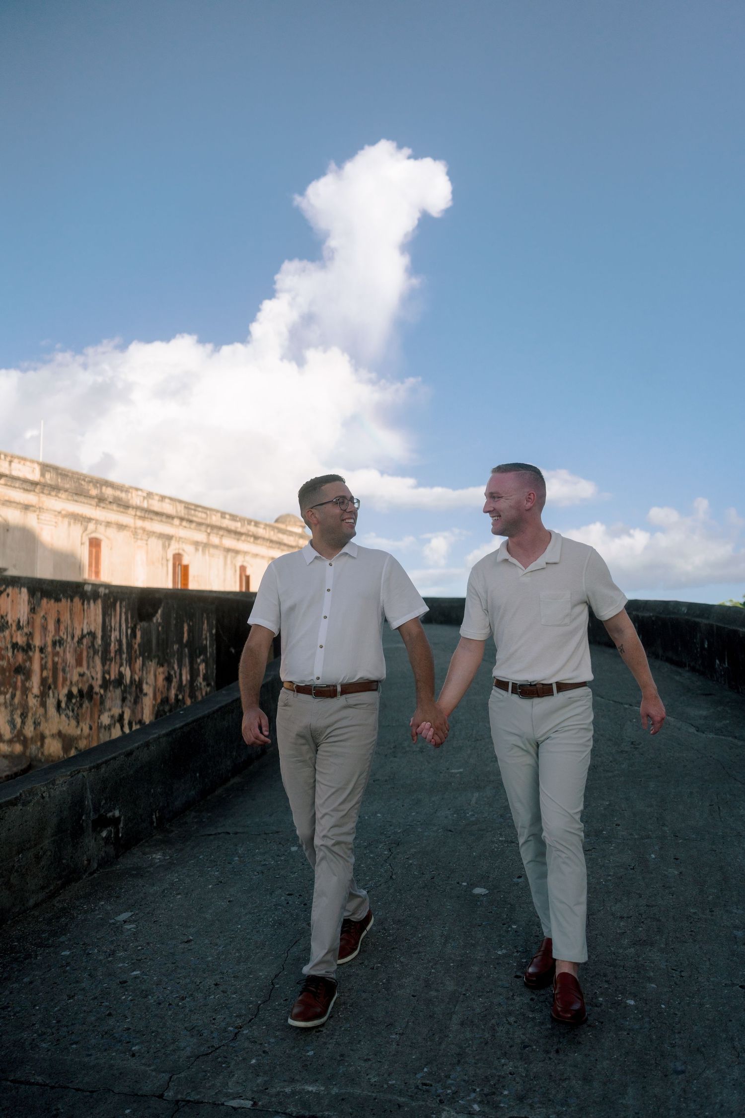 Two men in matching white shirts and khakis hold hands while walking on a historic stone walkway under a blue sky.