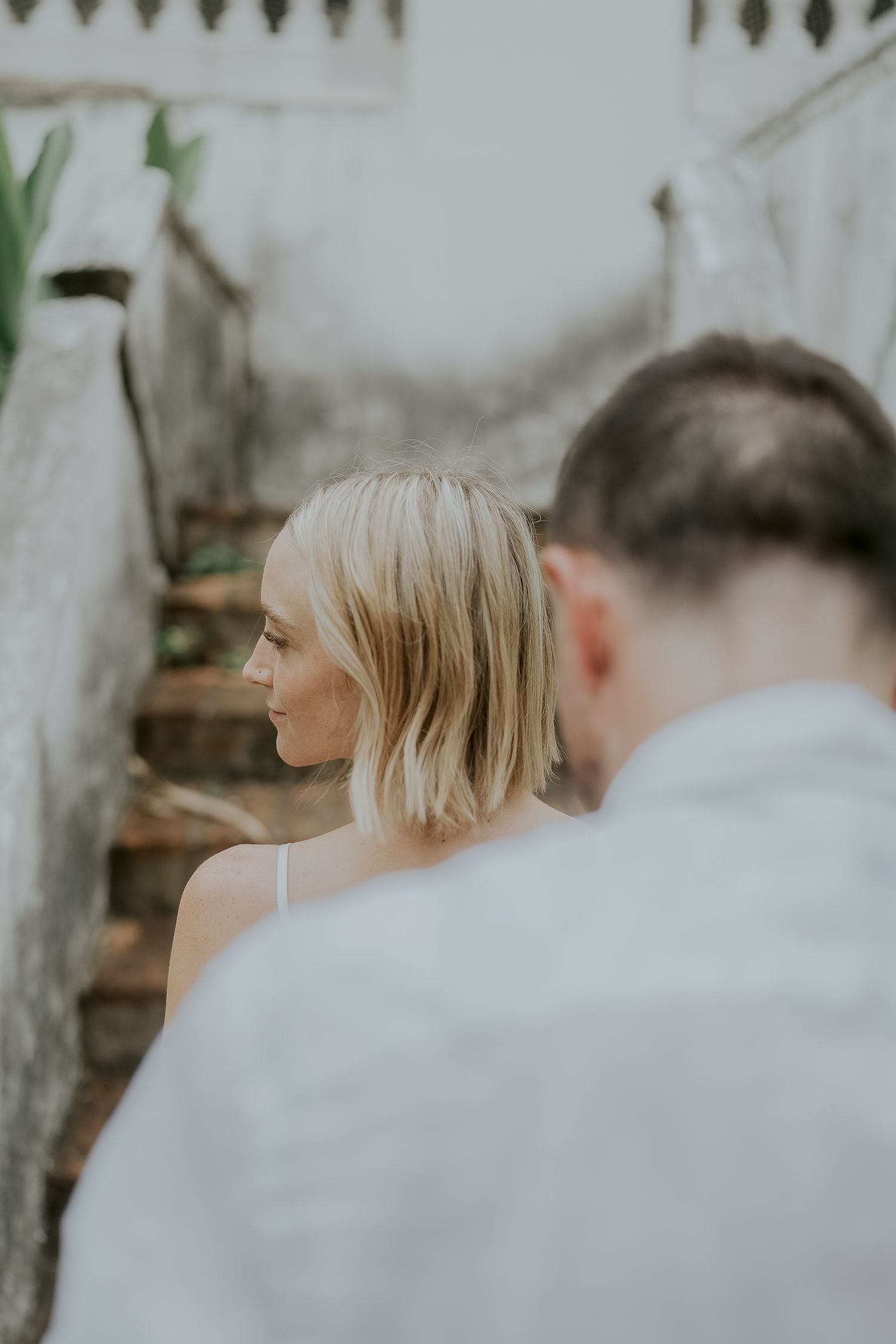 Blonde woman with short bob hair in a white dress stands near rustic stone stairs with a blurred man in foreground.