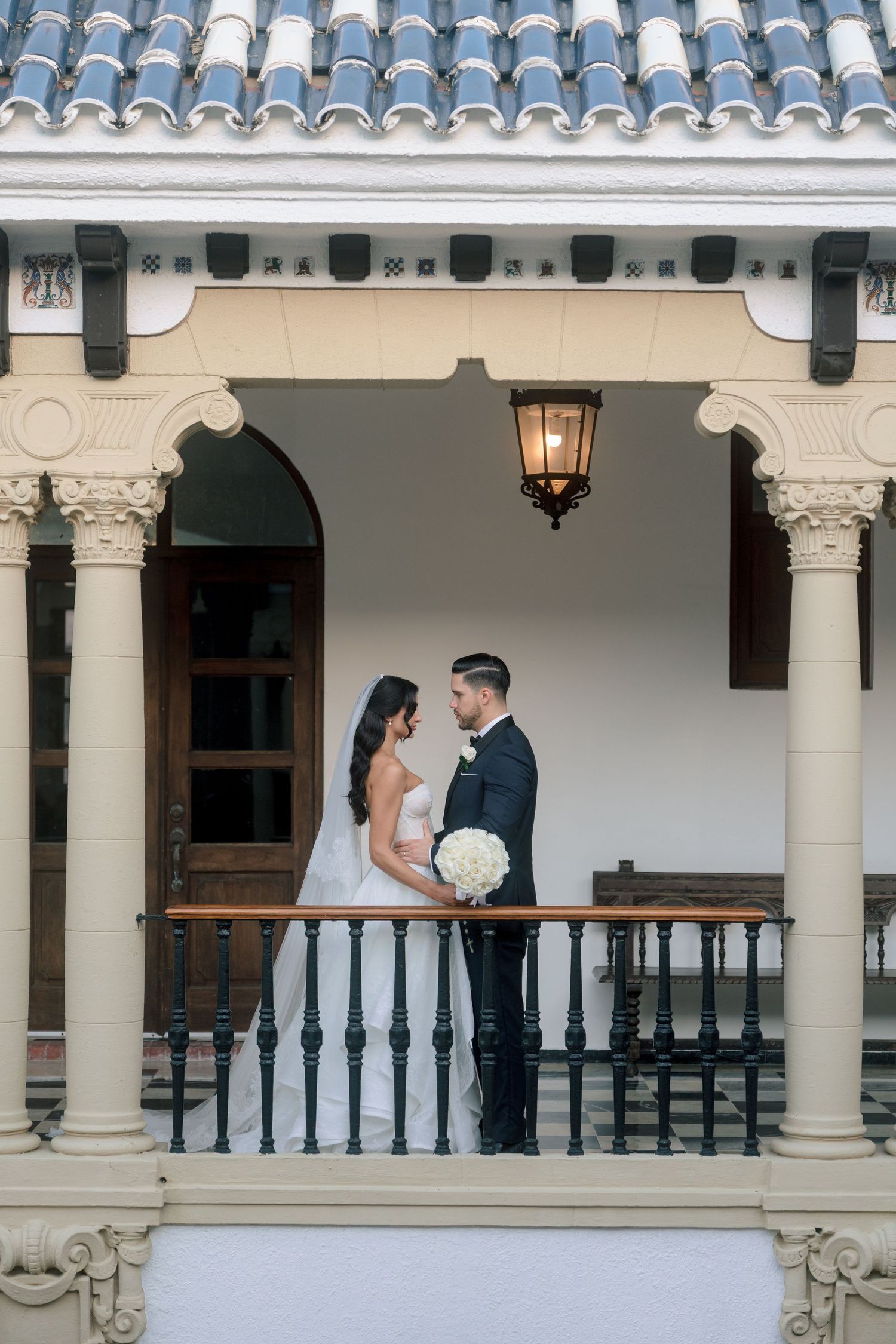 Bride and groom share a romantic moment on an elegant balcony with ornate columns and a lantern.