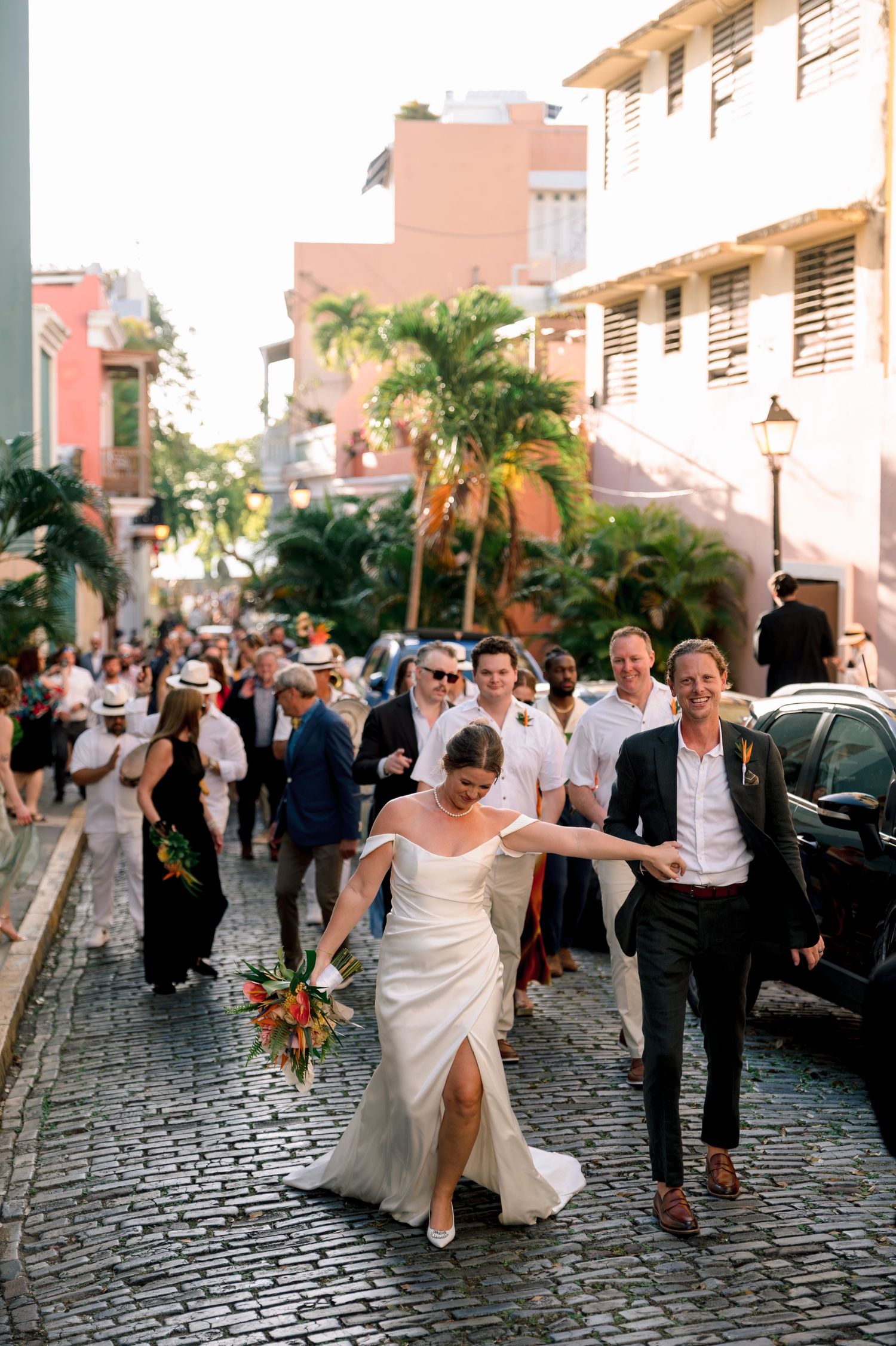 Bride and groom walking cobblestone street in Old San Juan with wedding guests following behind.